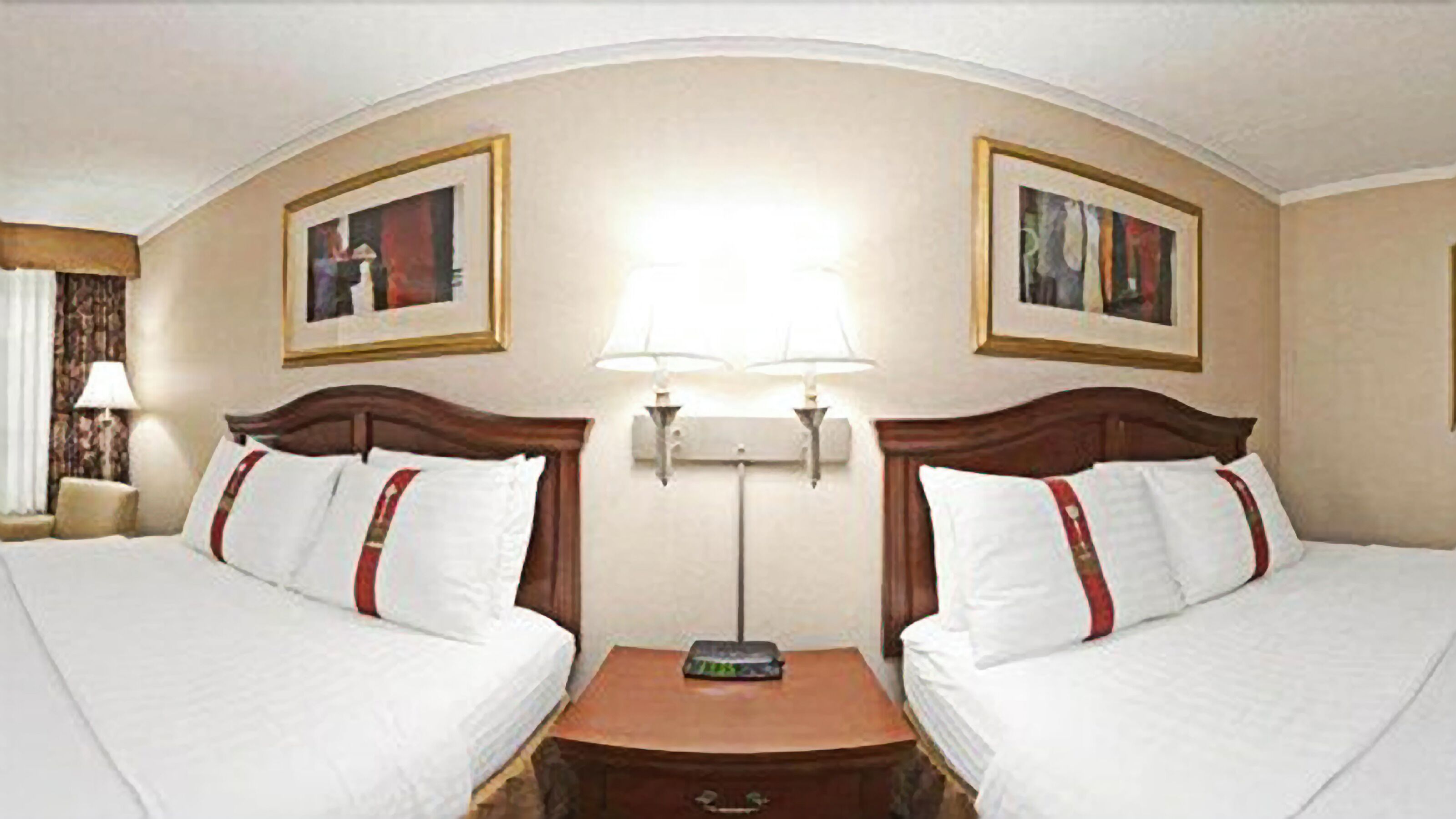 Habitación de hotel con dos camas, una mesita de noche, una lámpara y dos cuadros en las paredes.