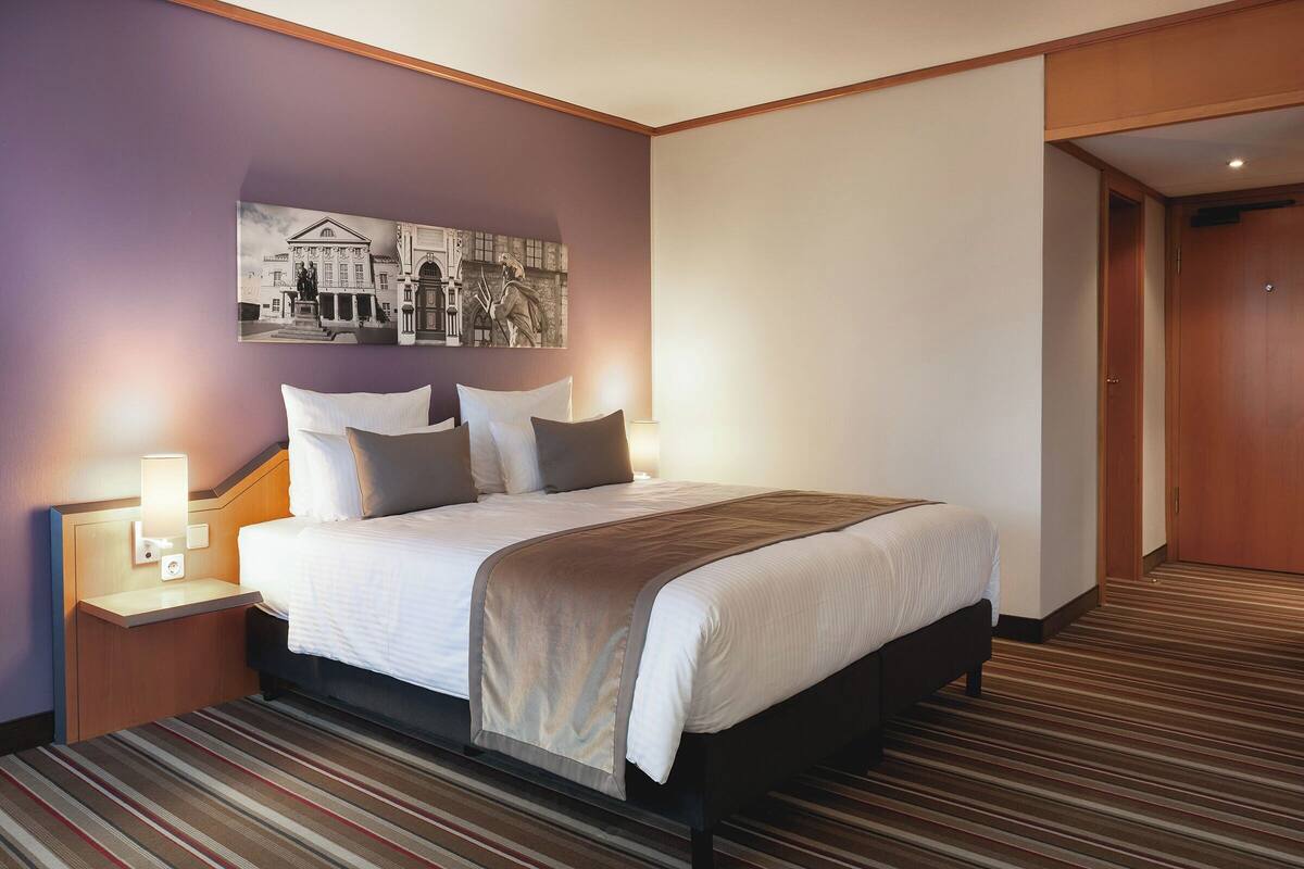 A hotel room with a large bed, a nightstand with a lamp, a wall-mounted picture, and a wooden door.