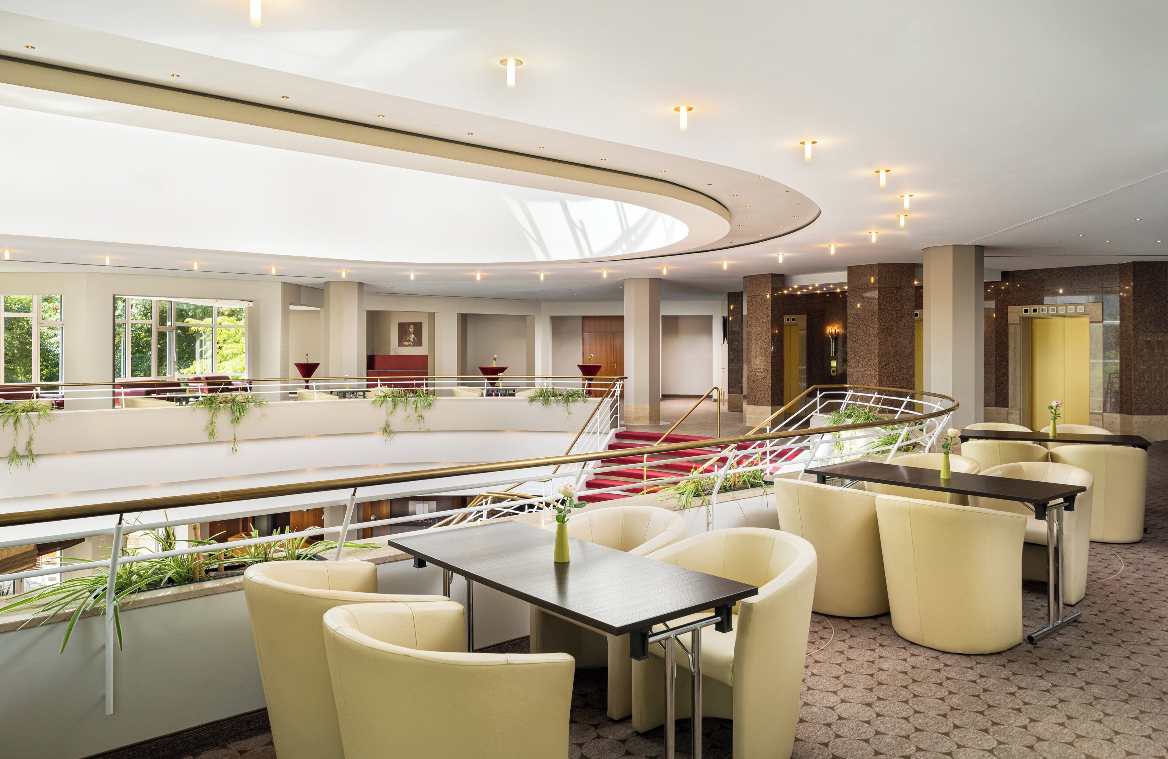 A modern hotel lobby with a spacious seating area, a central staircase, and a view of the outdoors.