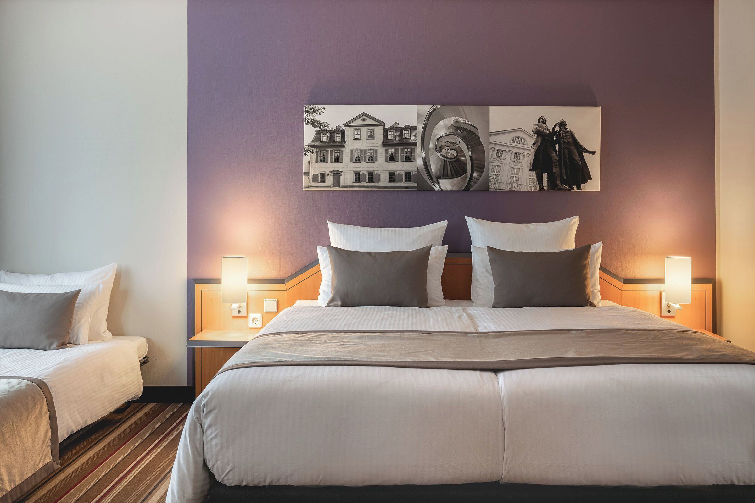 A hotel room with a large bed, two bedside tables, and a wall-mounted artwork depicting a building and two figures.