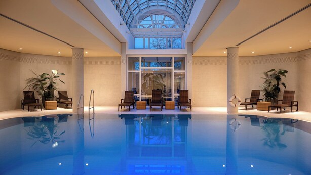 An indoor swimming pool with a glass ceiling, lounge chairs, and potted plants. - Leonardo Hotel Weimar (Weimar)