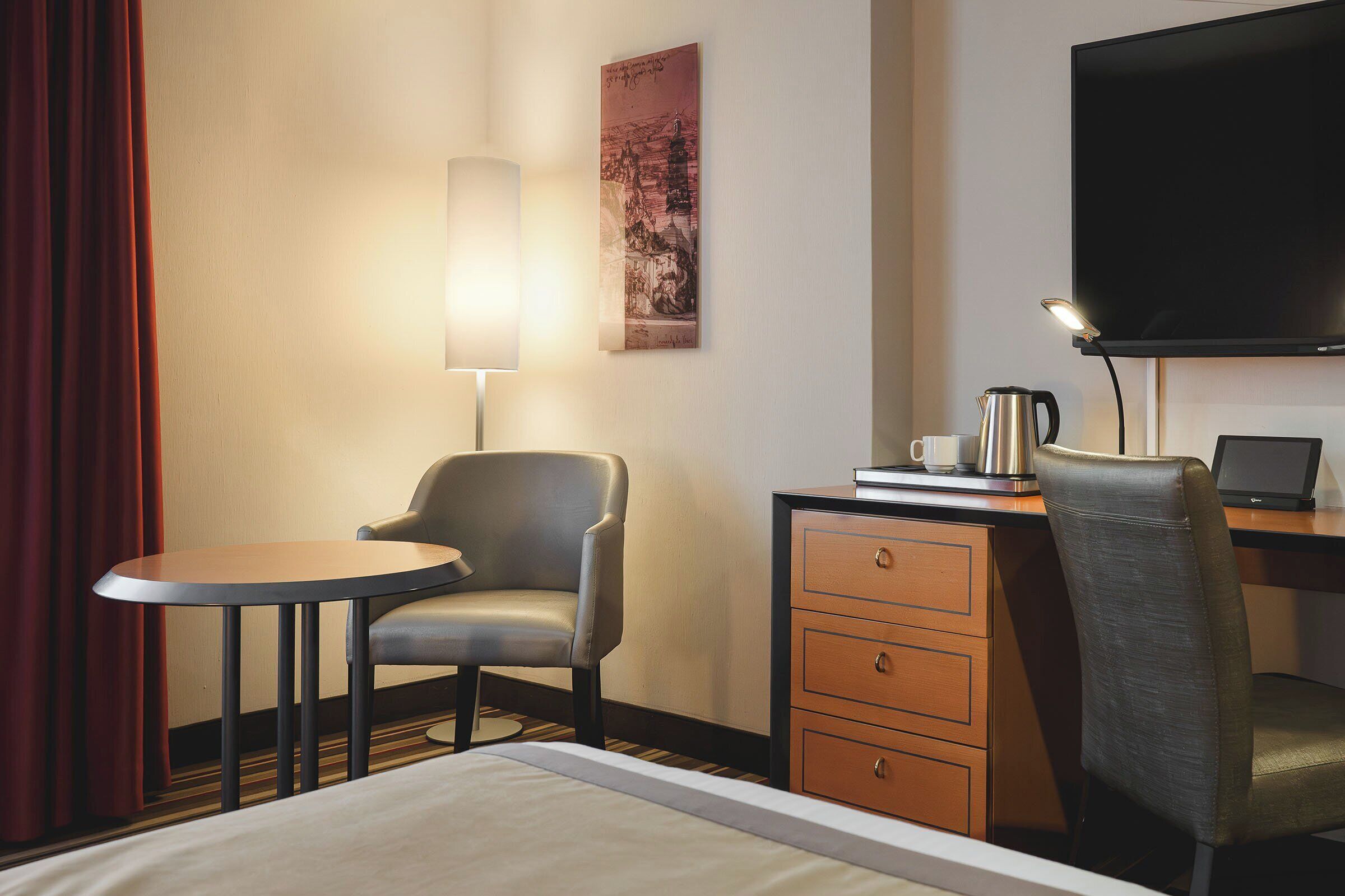 A hotel room with a bed, a desk with a laptop, a chair, a small table, a lamp, and a television.