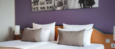 A double bed with white and beige bedding, a headboard with a wooden panel, and a wall-mounted artwork depicting a building and a statue.