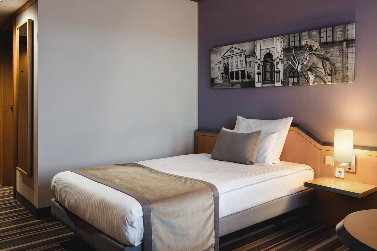 A hotel room with a bed, a nightstand with a lamp, a wall-mounted artwork, and a striped carpet.