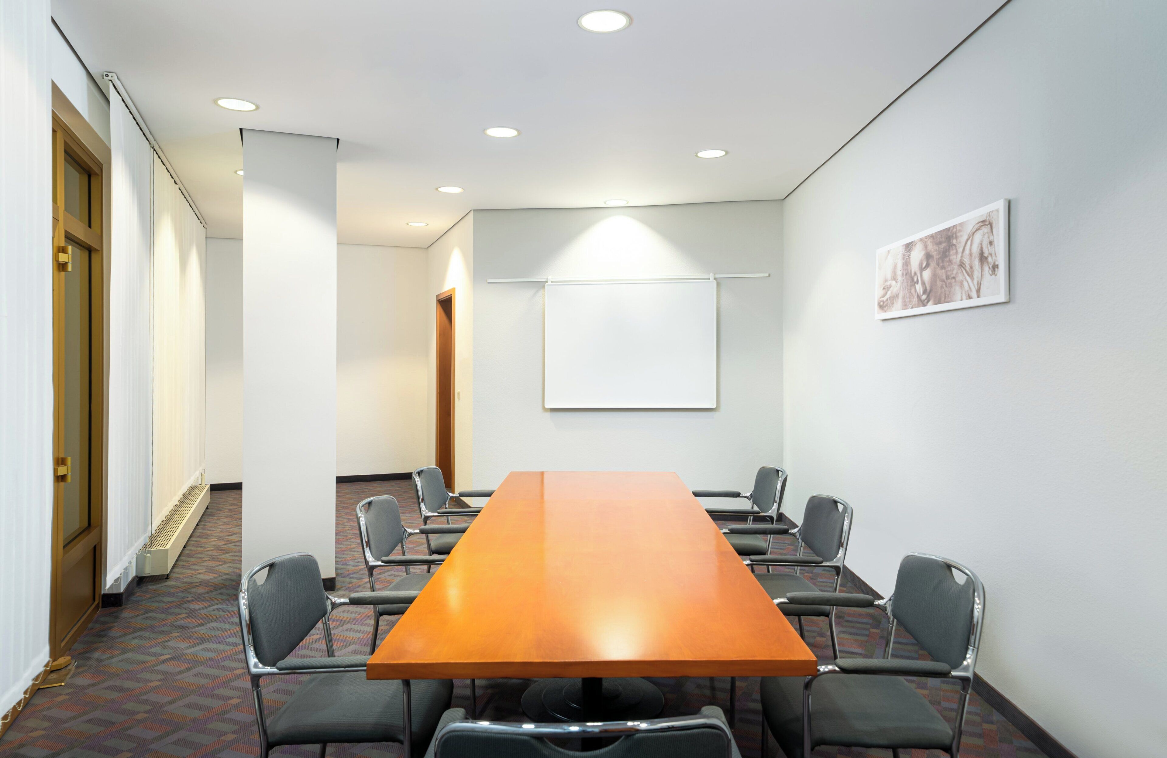 Ein Konferenzraum mit einem langen Tisch, sechs Stühlen, einer Projektionsleinwand und einer Tür.