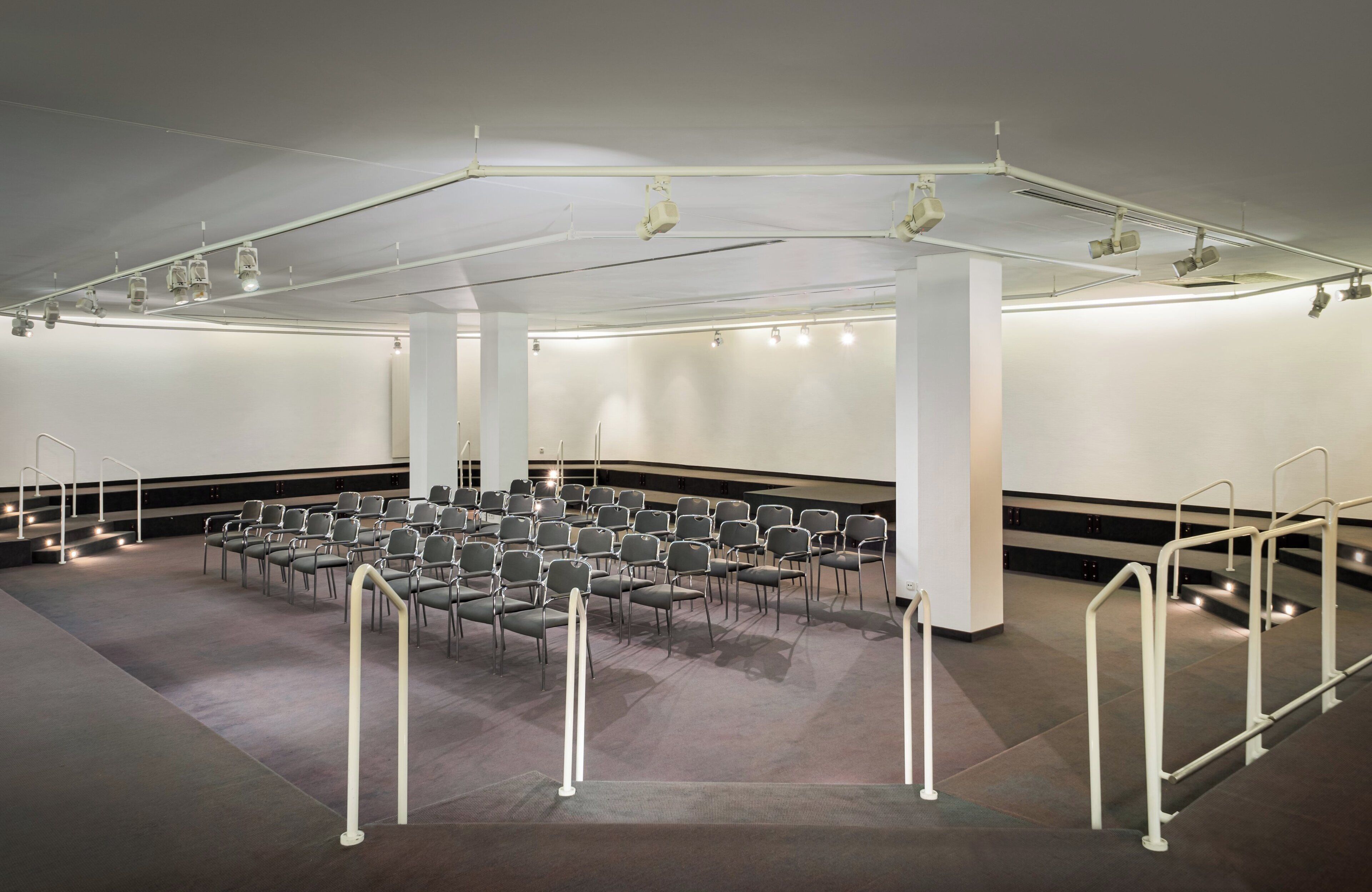 Ein Konferenzraum mit Reihen von Stühlen, einer Bühne inklusive Beleuchtung und einer hohen Decke.