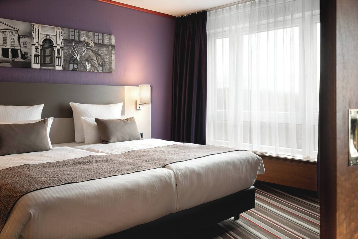 A hotel room with a large bed, a headboard with a cityscape painting, a window with sheer curtains, and a wooden floor.