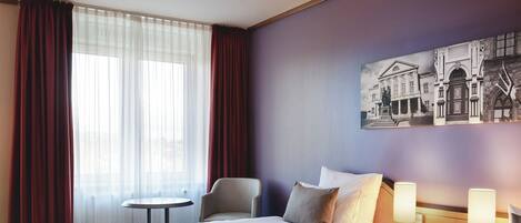 A hotel room with two beds, a small table, a chair, and a wall-mounted lamp.