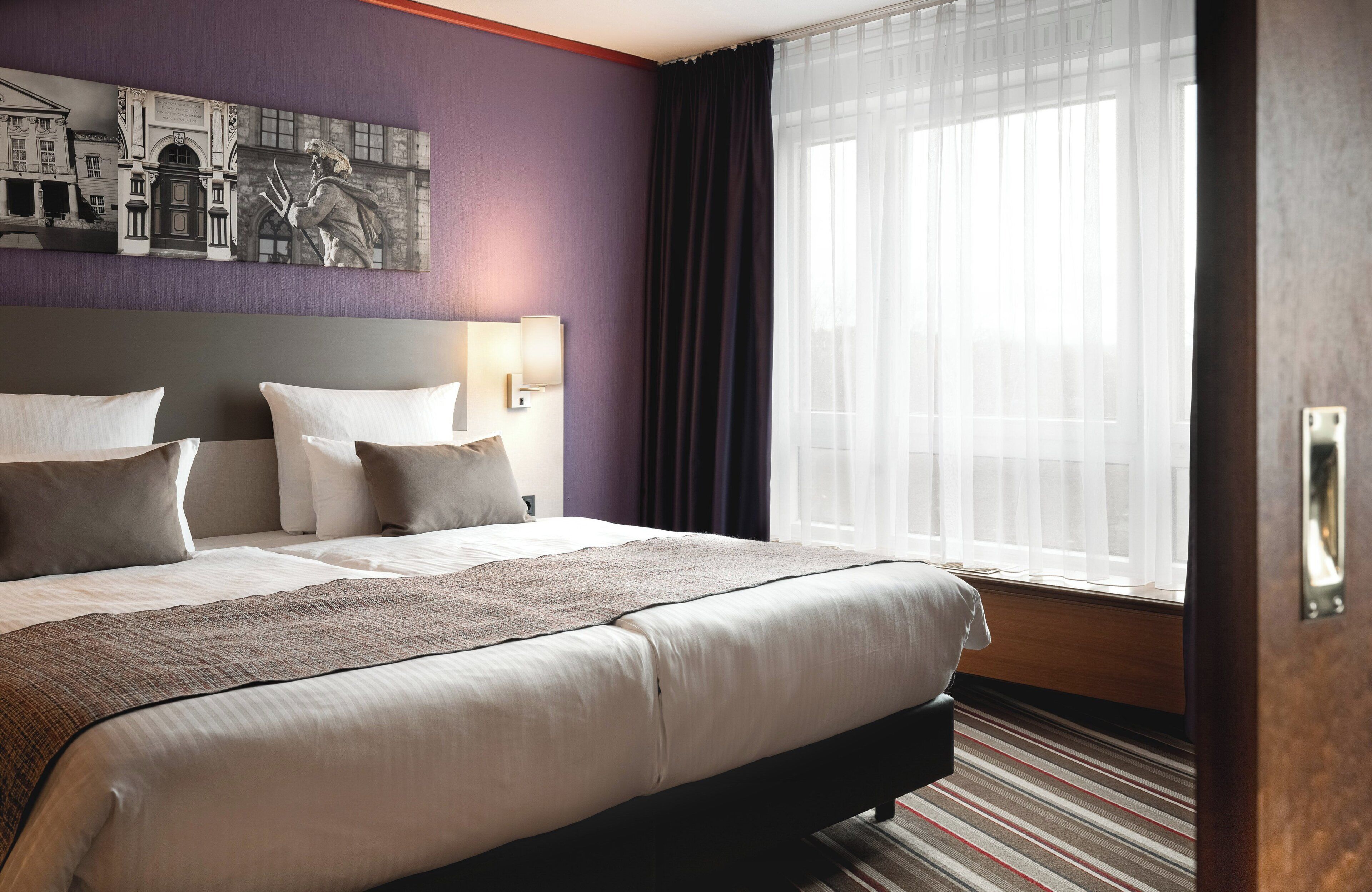 A hotel room with a large bed, a headboard with a cityscape painting, a window with sheer curtains, and a wooden floor.
