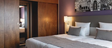 A hotel room with a large bed, a wooden wardrobe, a wall-mounted light, and a painting above the bed.