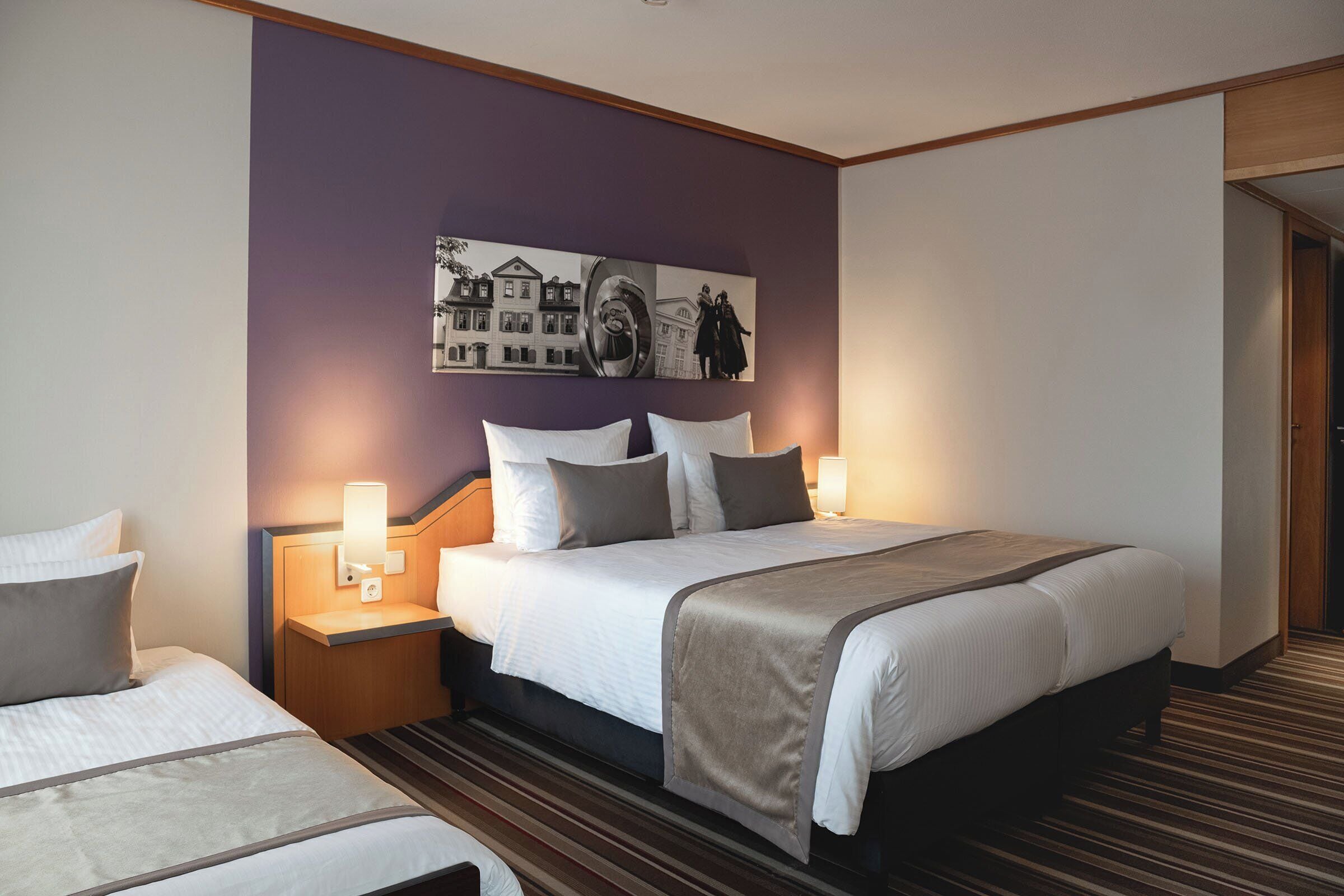 A hotel room with a large bed, a smaller bed, a nightstand with a lamp, and a wall-mounted artwork.