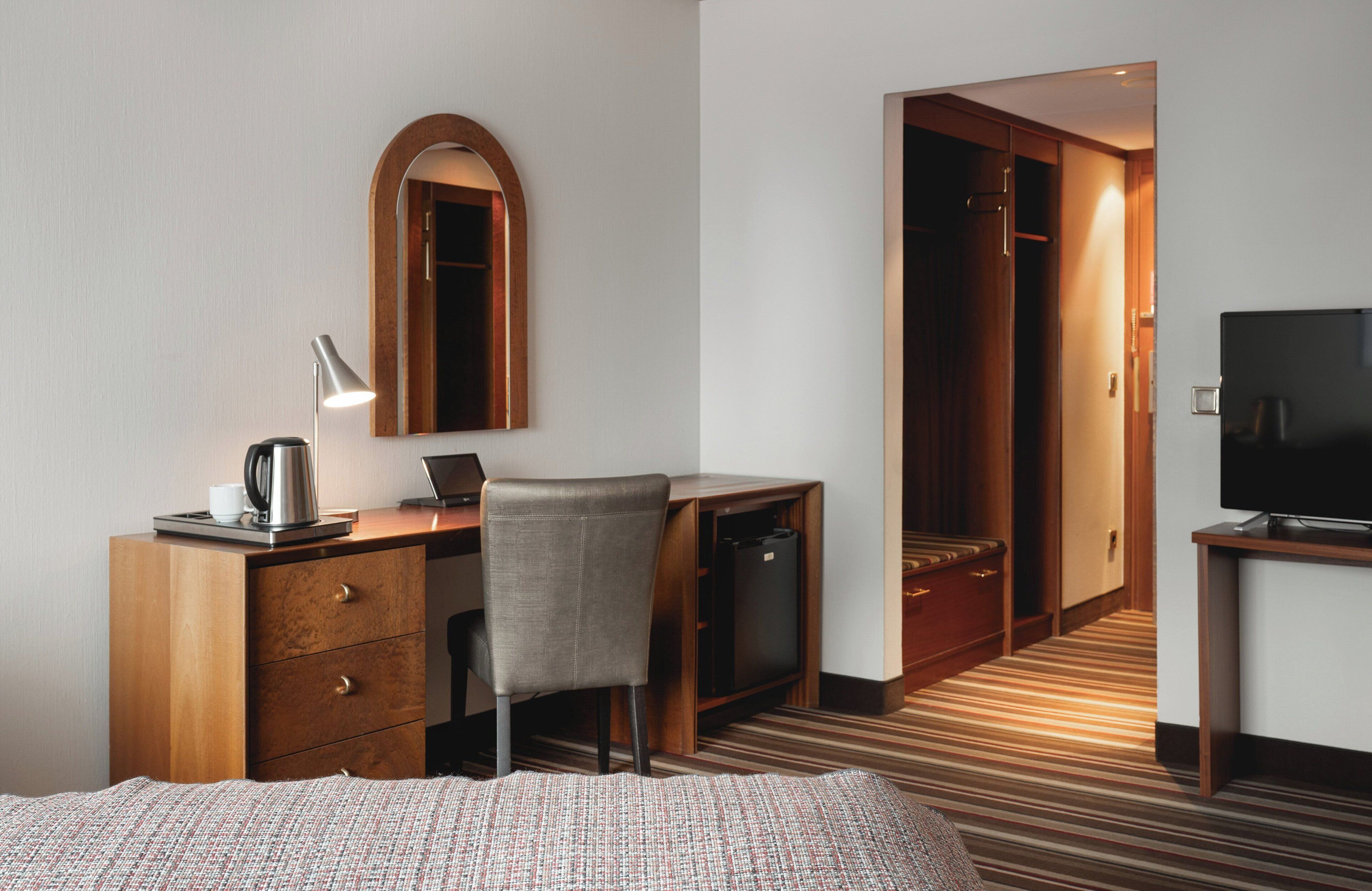 A hotel room with a bed, a desk with a chair, a television, a mirror, and a wardrobe.