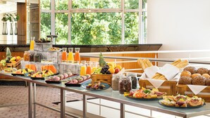 A buffet with various food items and beverages, a large window with a view of trees, and a modern interior design. - Leonardo Hotel Weimar (Weimar)