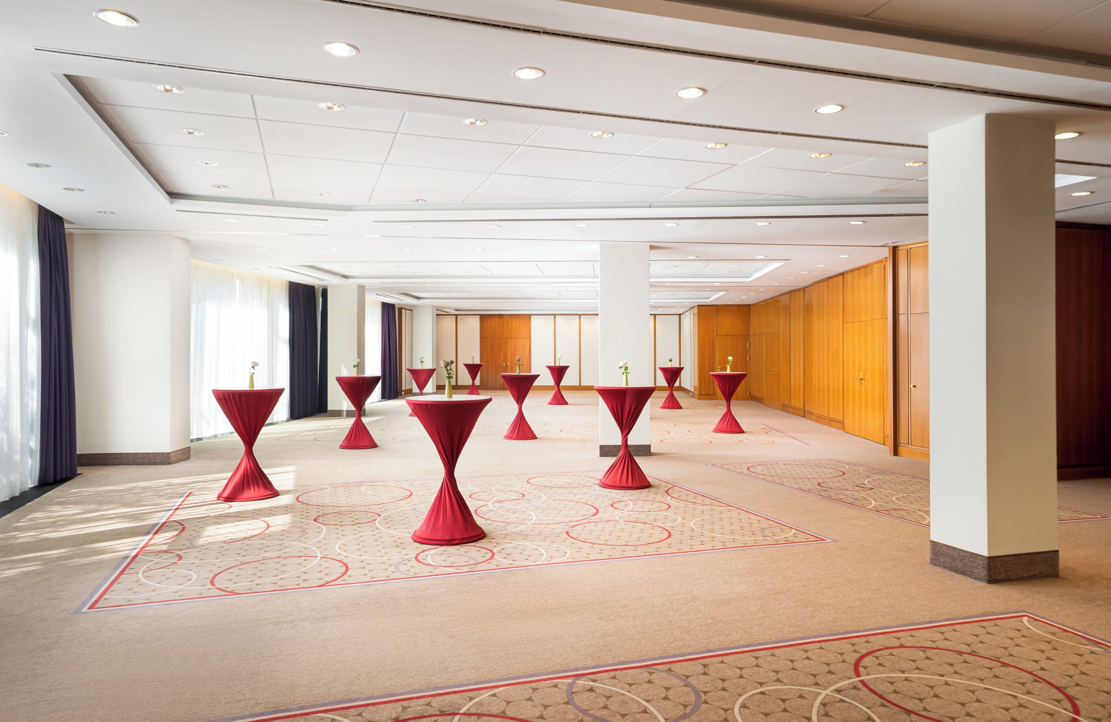 A spacious hotel corridor with multiple red tables and chairs, a patterned carpet, and wooden paneling on the walls.