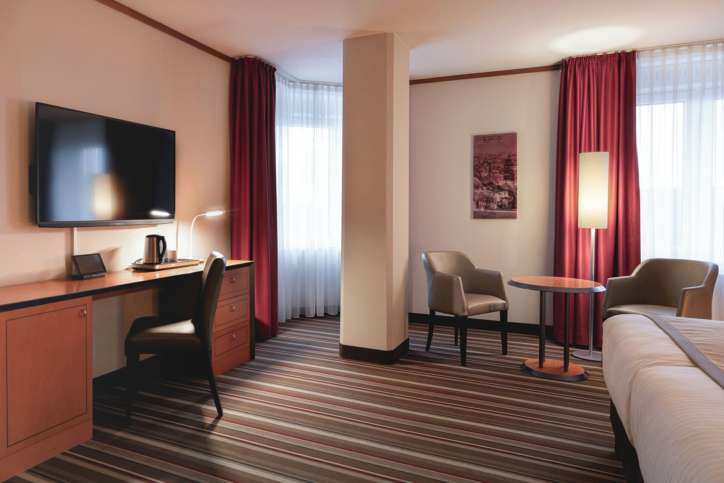 Habitación de hotel con cama, escritorio con computadora portátil, silla, mesa pequeña, televisor y lámpara.