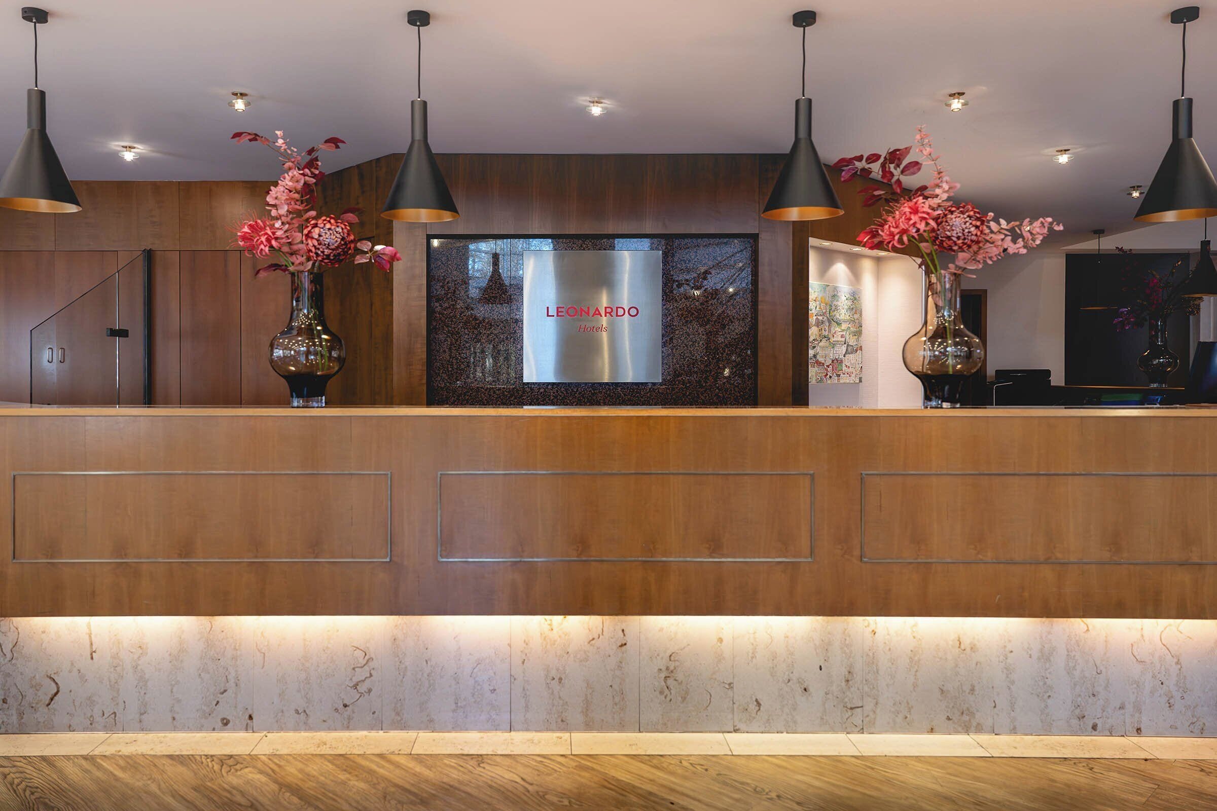 A hotel reception desk with a digital display, wooden paneling, and two vases with flowers.