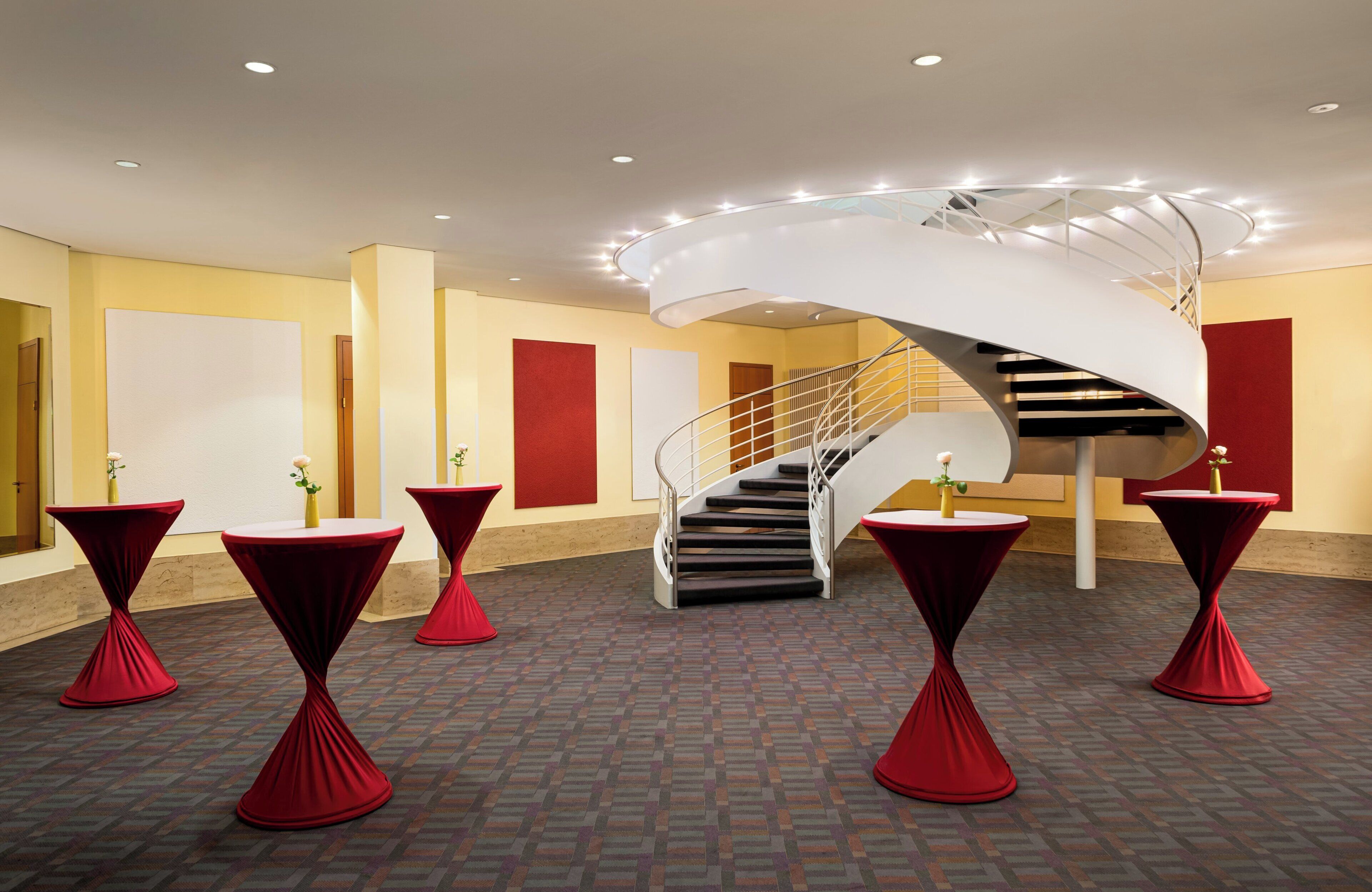 Eine moderne Hotelhalle mit einer Wendeltreppe, roten Tischen mit weißen Oberflächen und einem Teppichboden mit Muster.