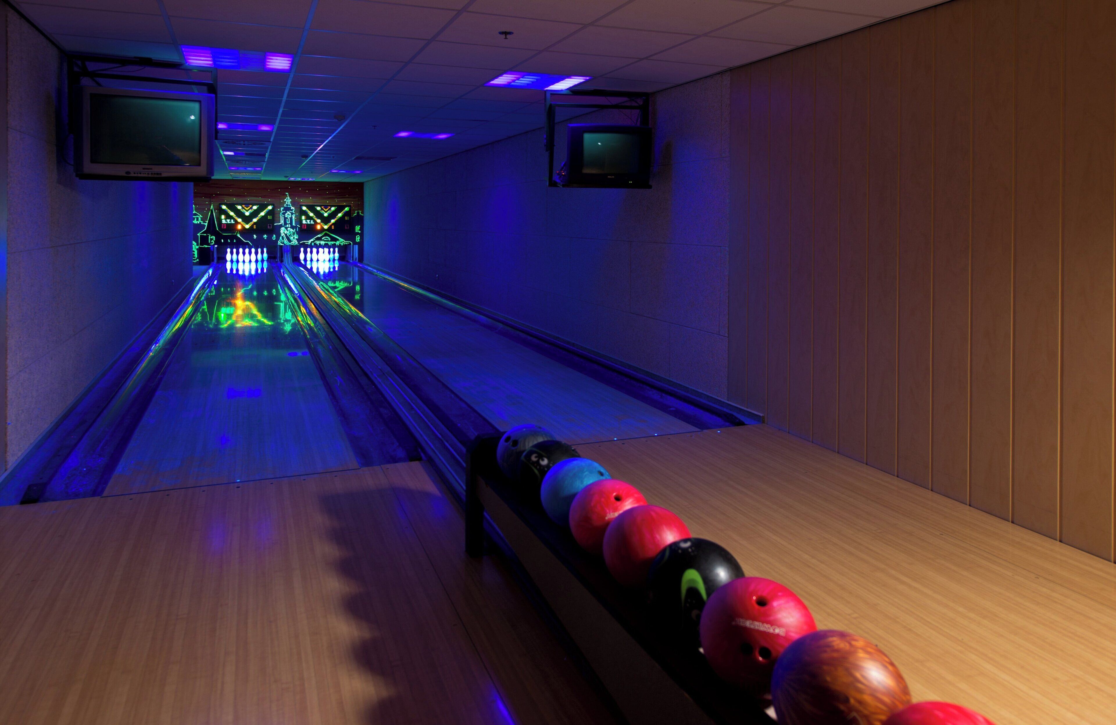 Une salle de bowling avec un plancher en bois, une rangée de quilles et deux téléviseurs suspendus au plafond.