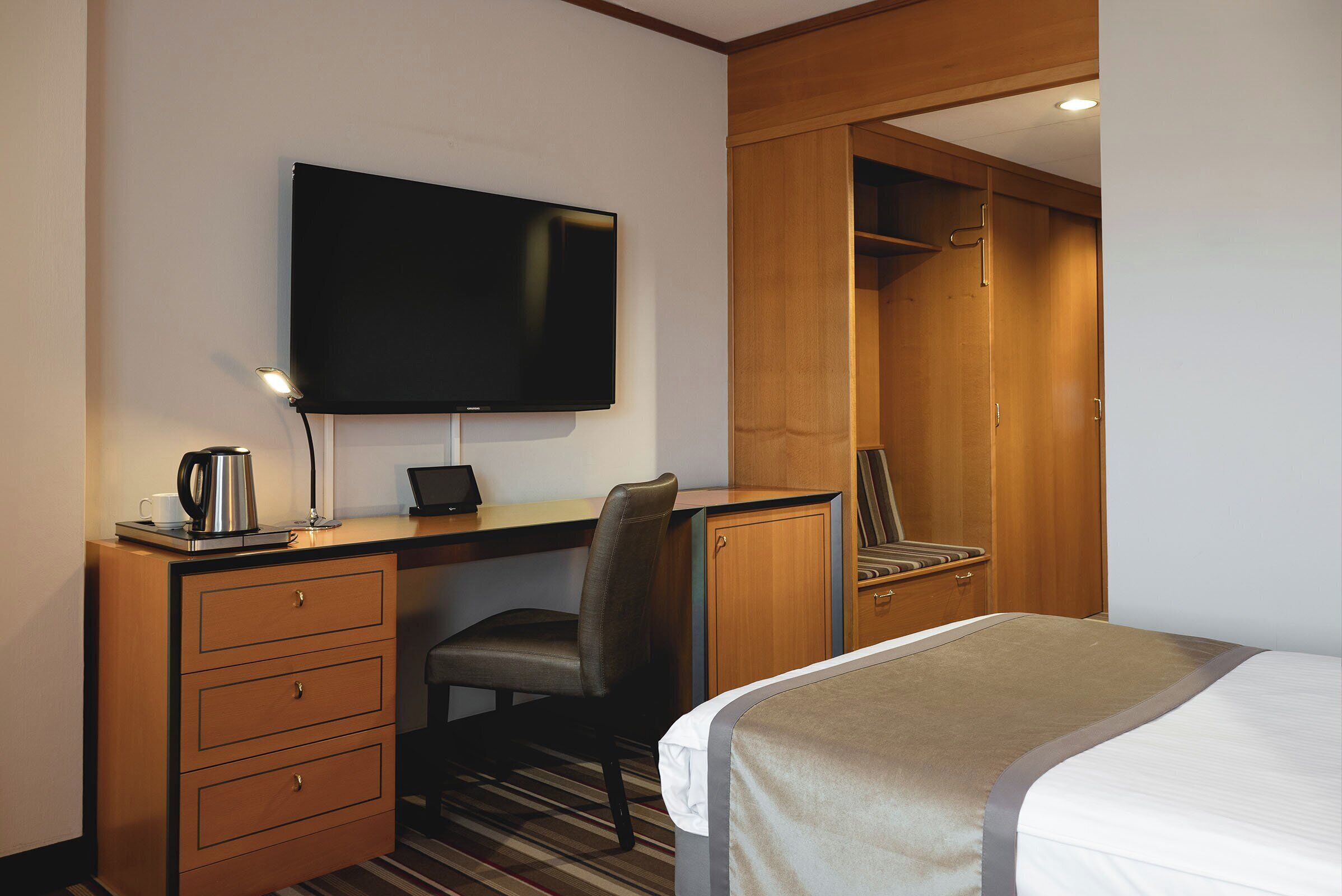 A hotel room with a bed, a desk with a computer, a chair, a television, and a wardrobe.