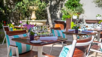Un espacio de comedor al aire libre con mesas dispuestas para dos, sillas con cojines a rayas y plantas en macetas.