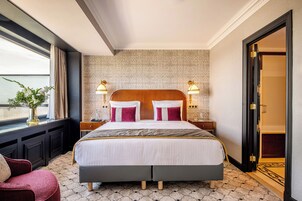 Ein Hotelzimmer mit einem großen Bett, zwei Nachttischlampen, einem Nachttisch, einem Fenster mit Vorhängen und einer Tür zu einem anderen Raum.