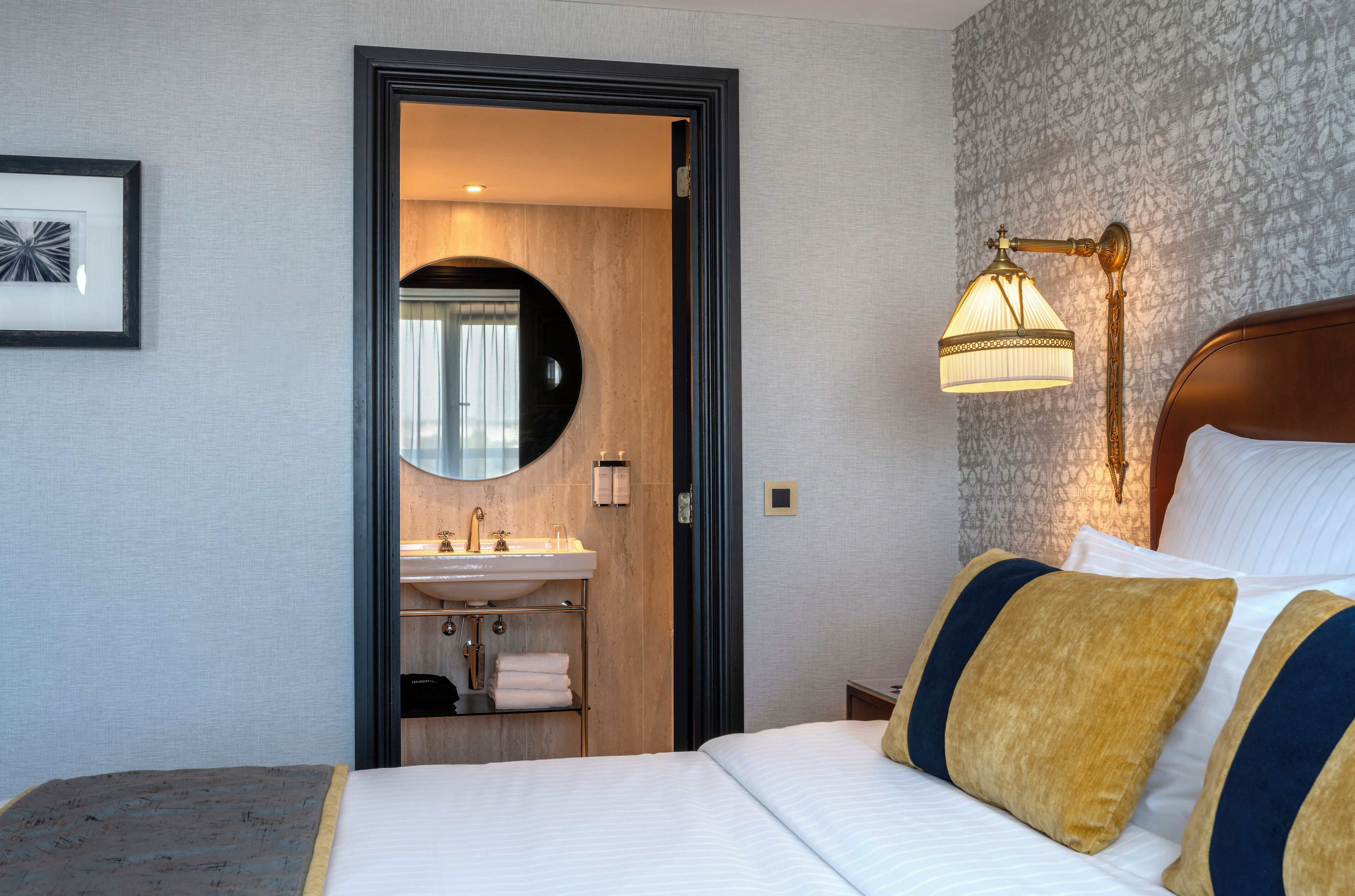 Een hotelkamer met een bed, een bedlampje, een spiegel en een badkamer met een wastafel en handdoekhouder.