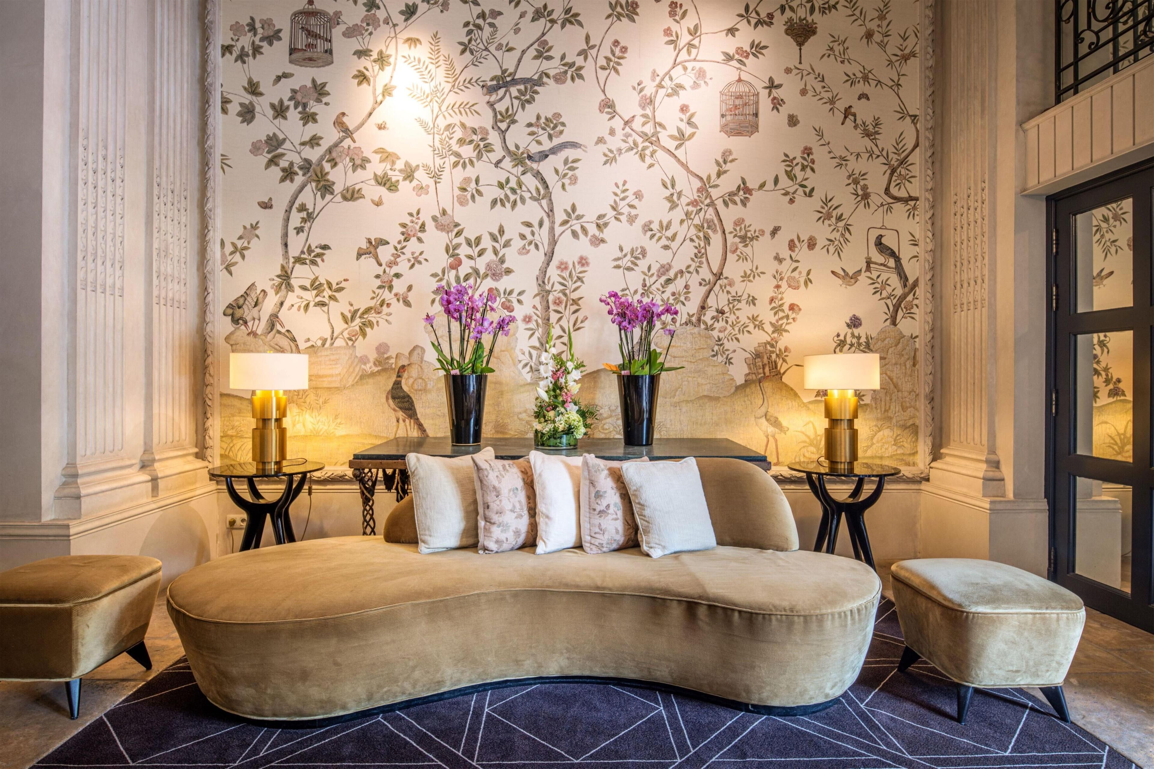 A room with a curved sofa, two lamps, and a floral wallpaper.