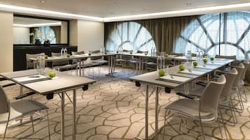 Un salón de conferencias con varias mesas y sillas, equipado con botellas de agua y manzanas, y un ventanal con un diseño geométrico.