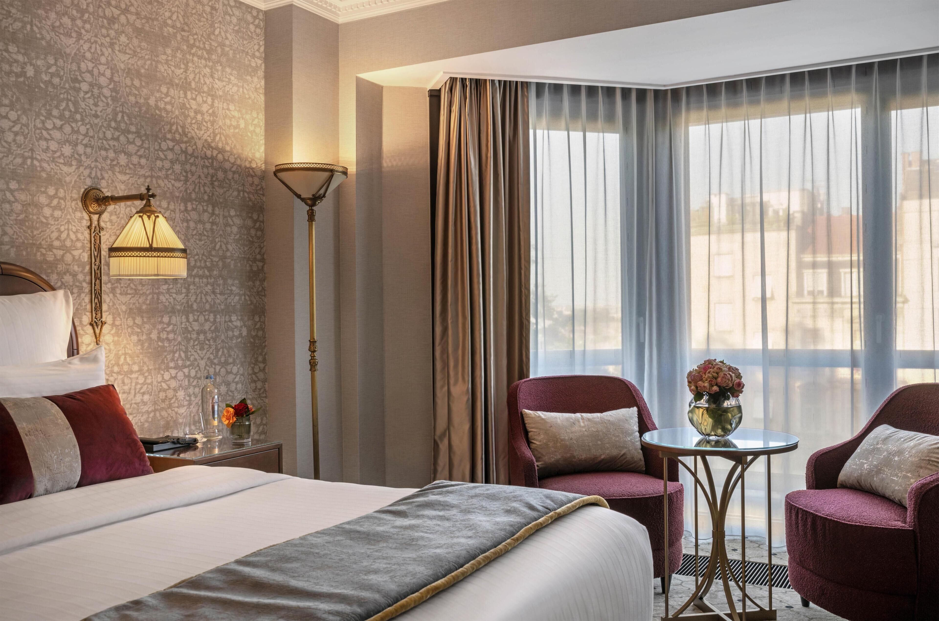 Habitación de hotel con cama, dos sillones, una mesita con un florero y una lámpara de pie.