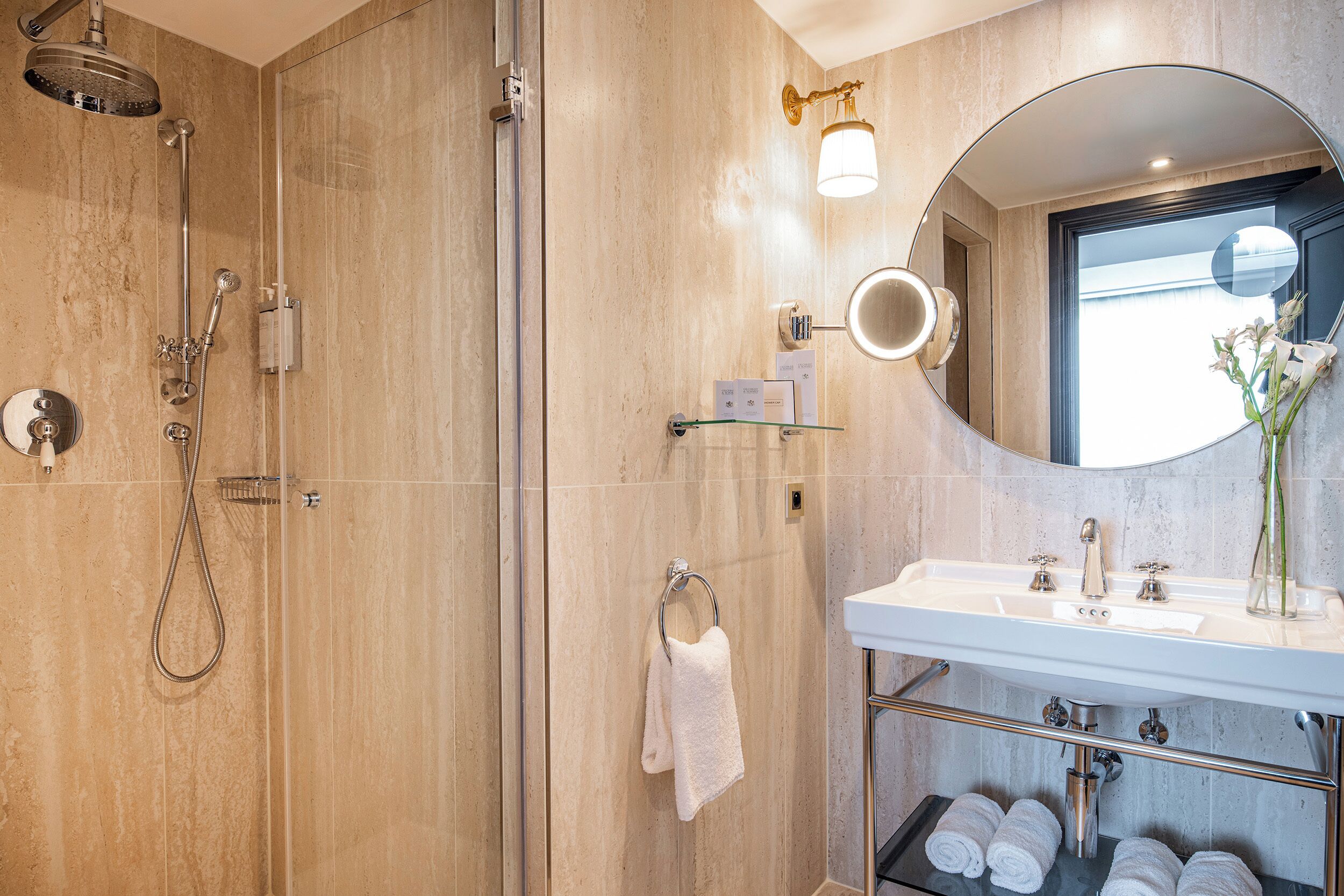 Un baño moderno con ducha de vidrio, un lavamanos blanco con espejo y toallas ordenadas en un estante.