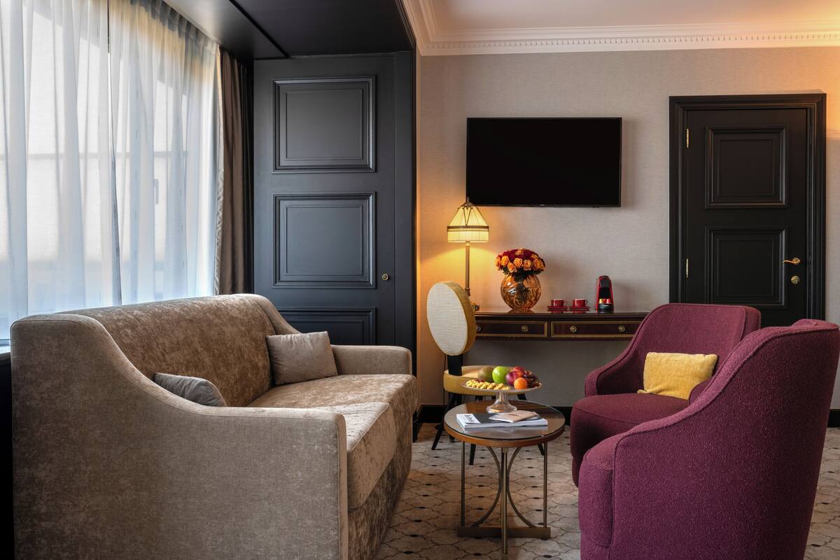 A hotel room with a sofa, armchair, small table with fruit, a lamp, and a TV on the wall.