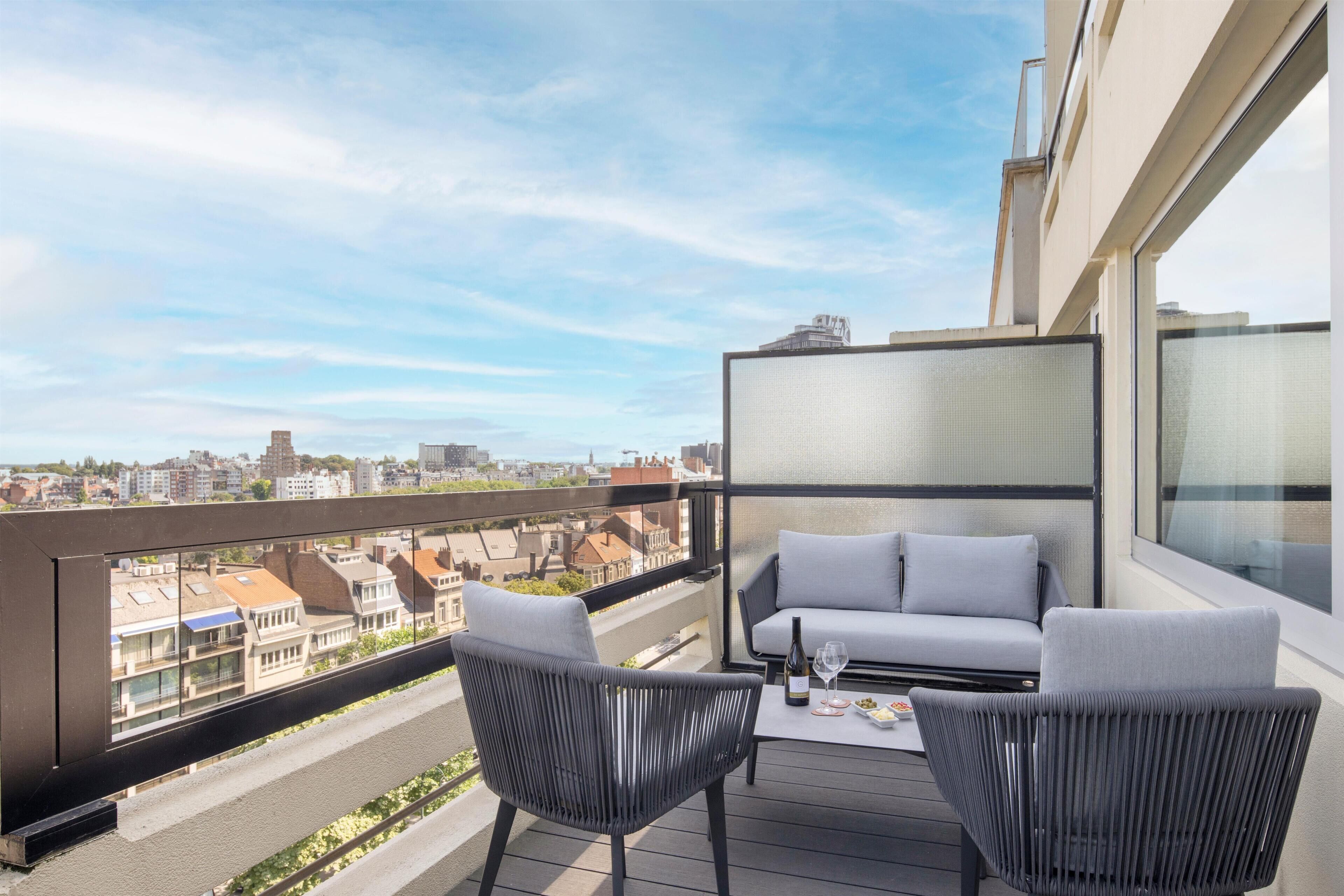 A balcony with a sofa and chairs, a table with a bottle and glasses, and a city view.