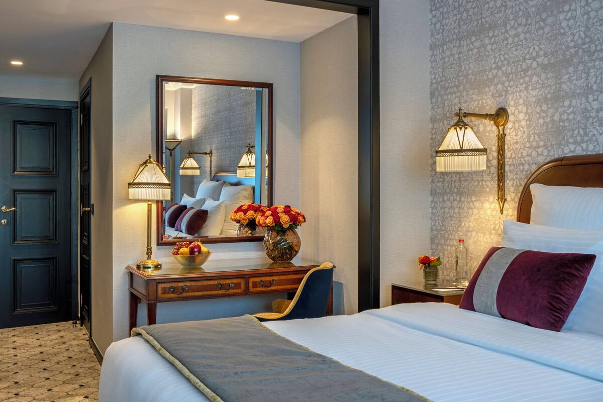 A hotel room with a bed, a desk with a vase of flowers, a mirror, and a nightstand with a lamp.