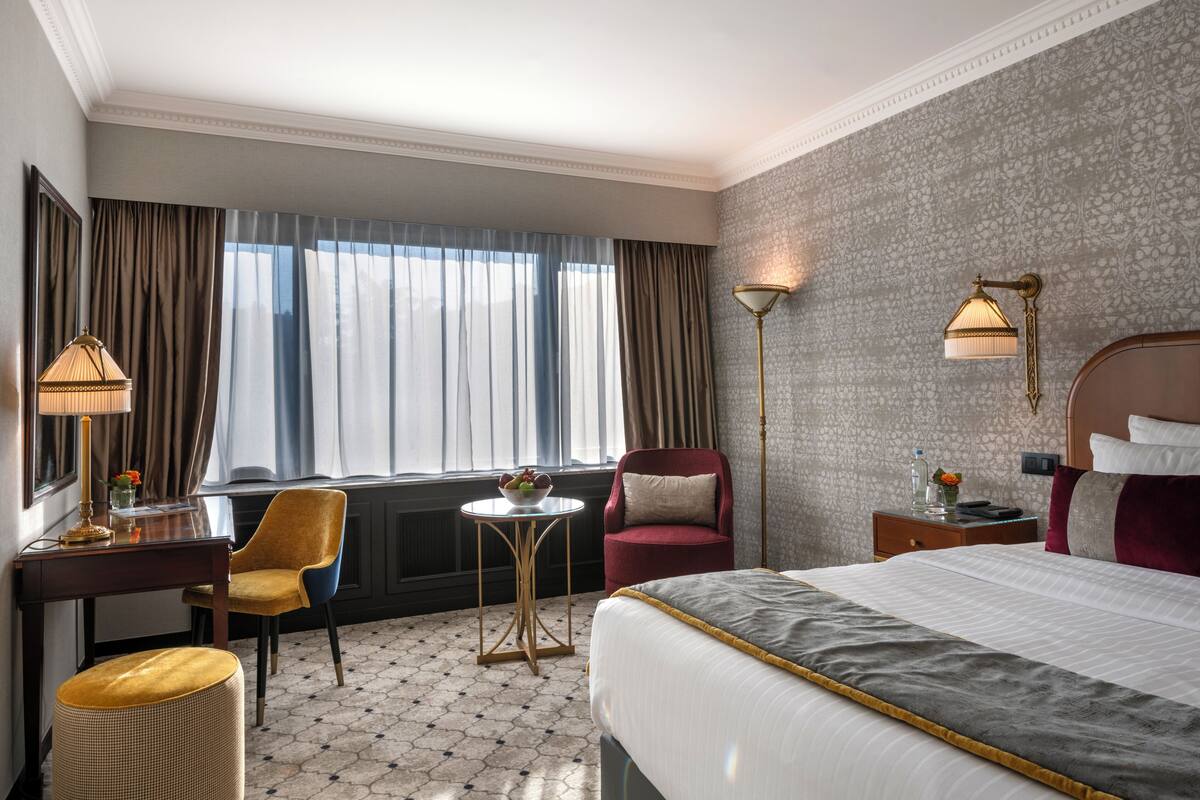 A hotel room with a bed, a desk, a chair, a lamp, a nightstand, and a window with curtains.