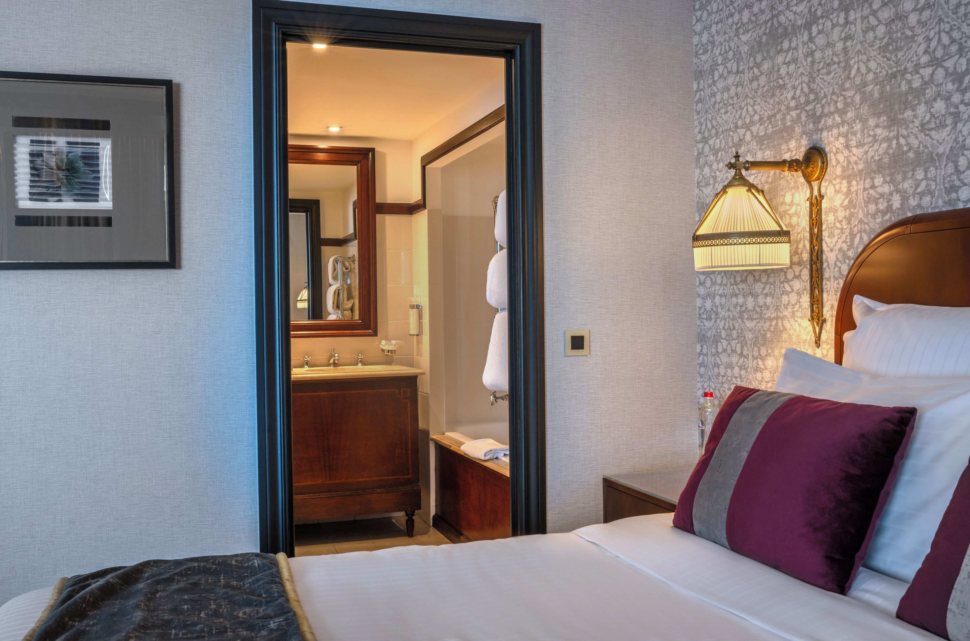 A hotel room with a bed, a nightstand, a lamp, a bathroom visible through an open door, and a framed picture on the wall.