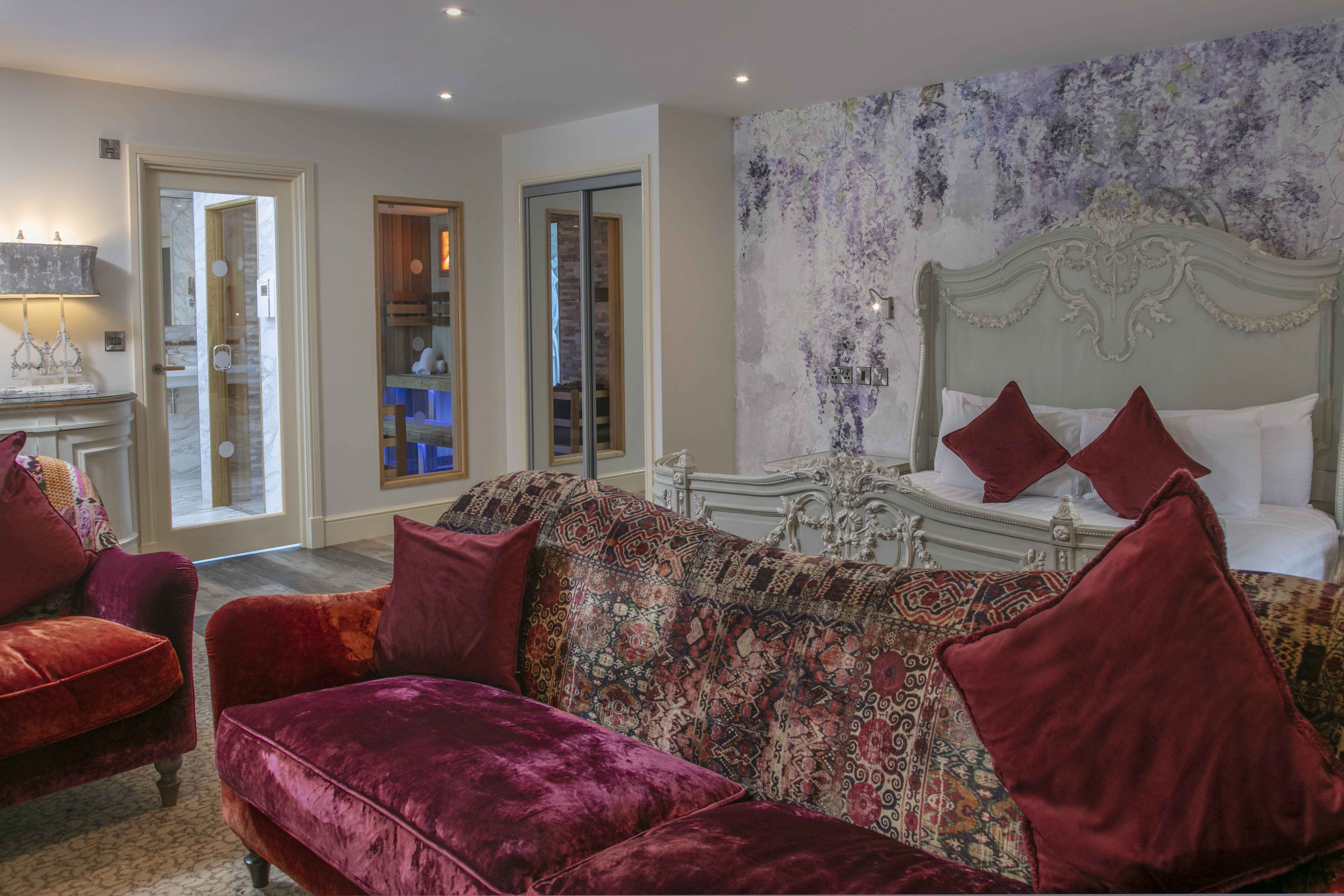 A room with a patterned sofa, a bed with red pillows, and a wall with a floral tapestry.