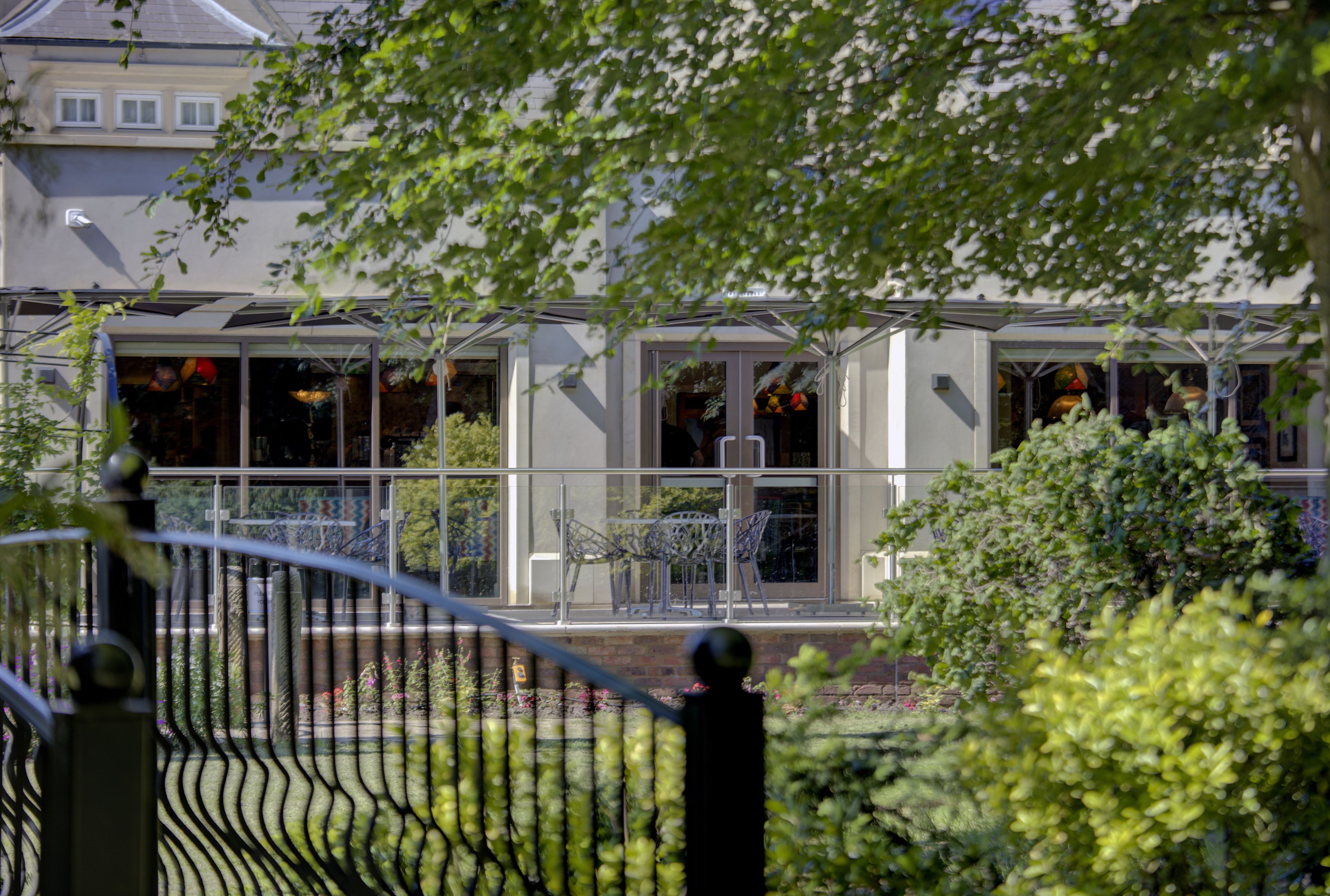 A conservatory with glass walls and metal chairs, surrounded by greenery and a fence.