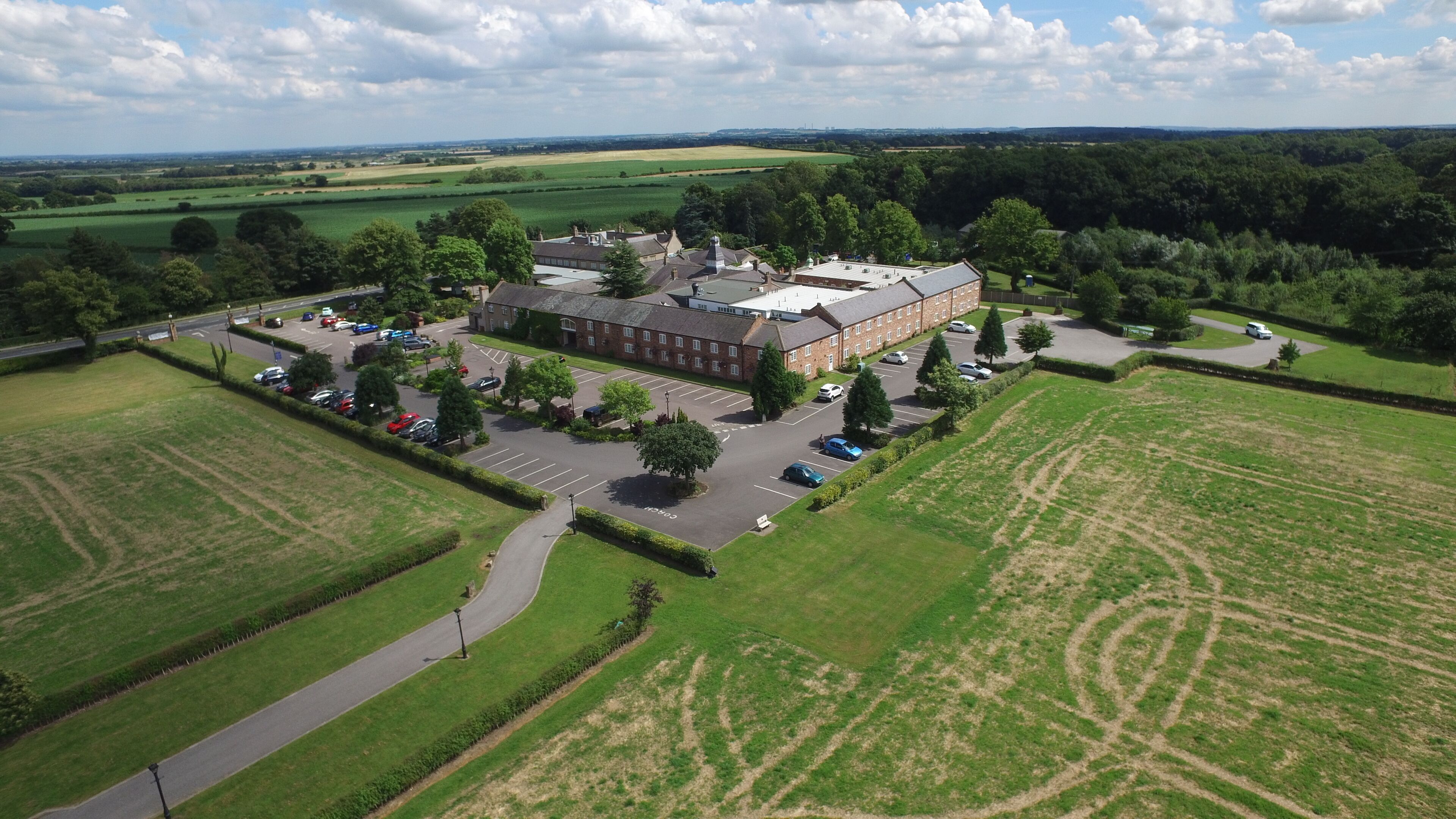 An aerial view of a large building with a parking lot, surrounded by green fields and trees.