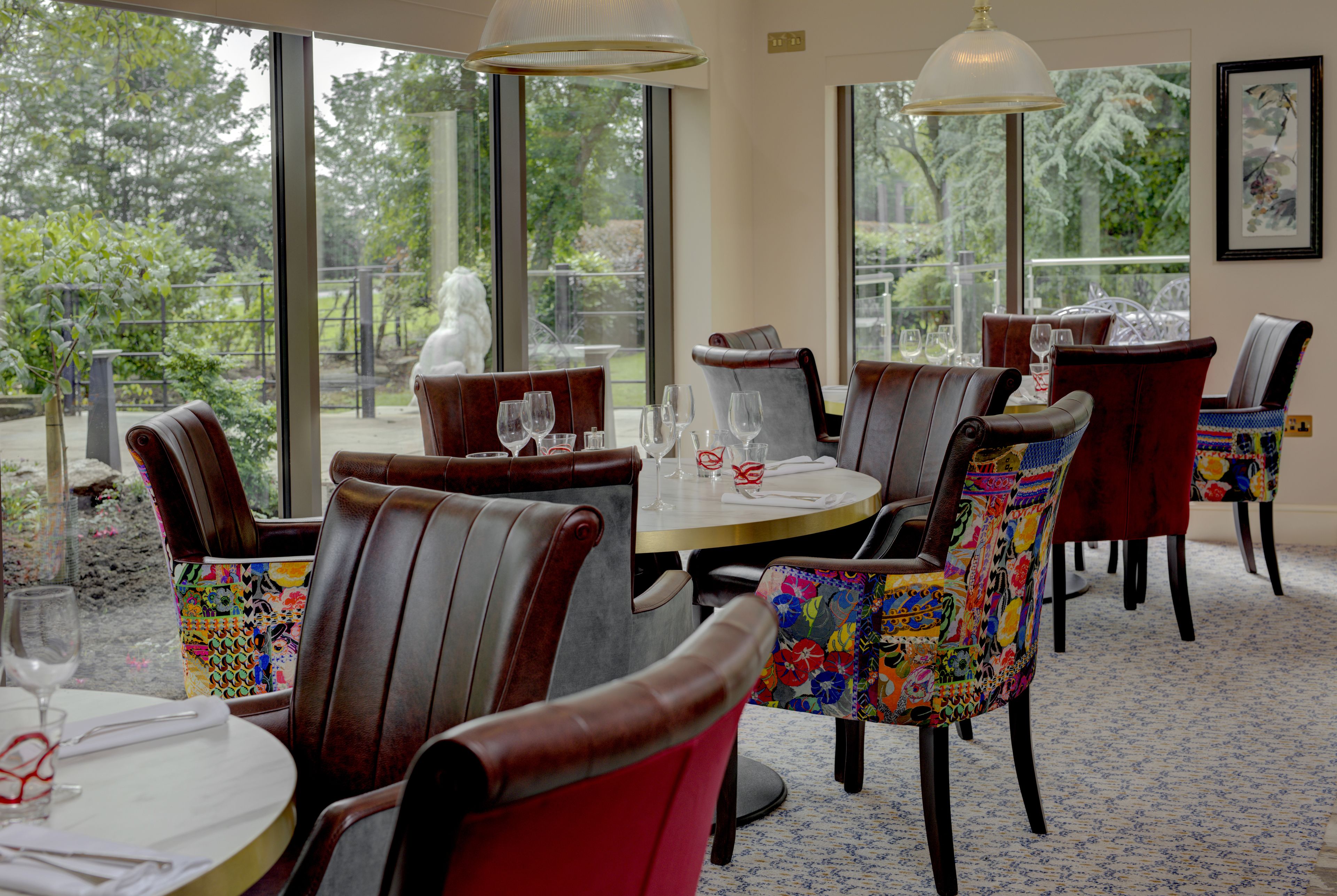 A restaurant with round tables and chairs, large windows with a view of greenery, and a framed picture on the wall.