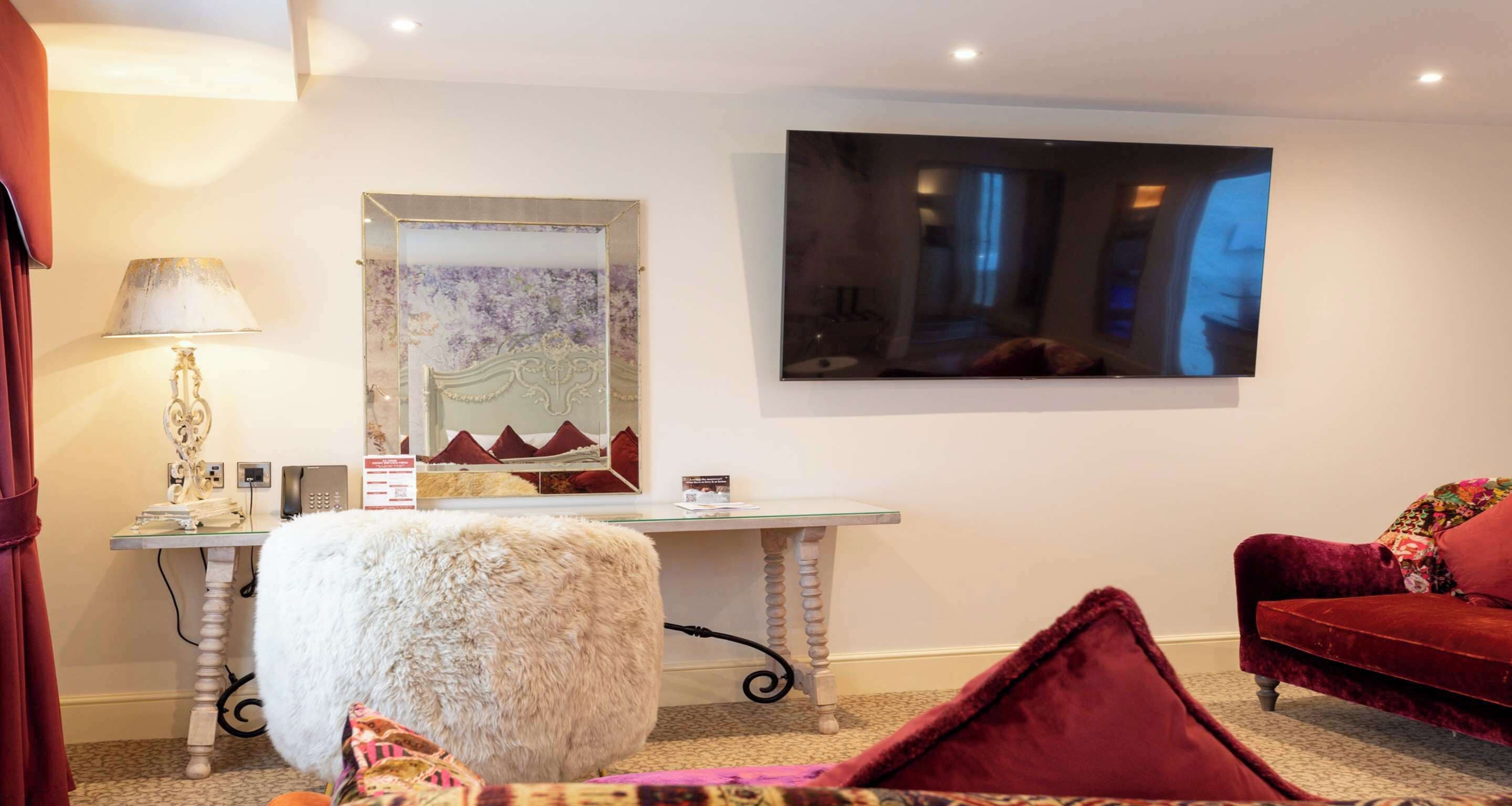 A room with a white tufted ottoman, a red velvet sofa, a glass desk with a mirror, and a large window.