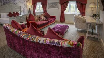 A room with a large, plush, purple sofa, a white bed with red pillows, a small round table, and a mirror with a lamp.