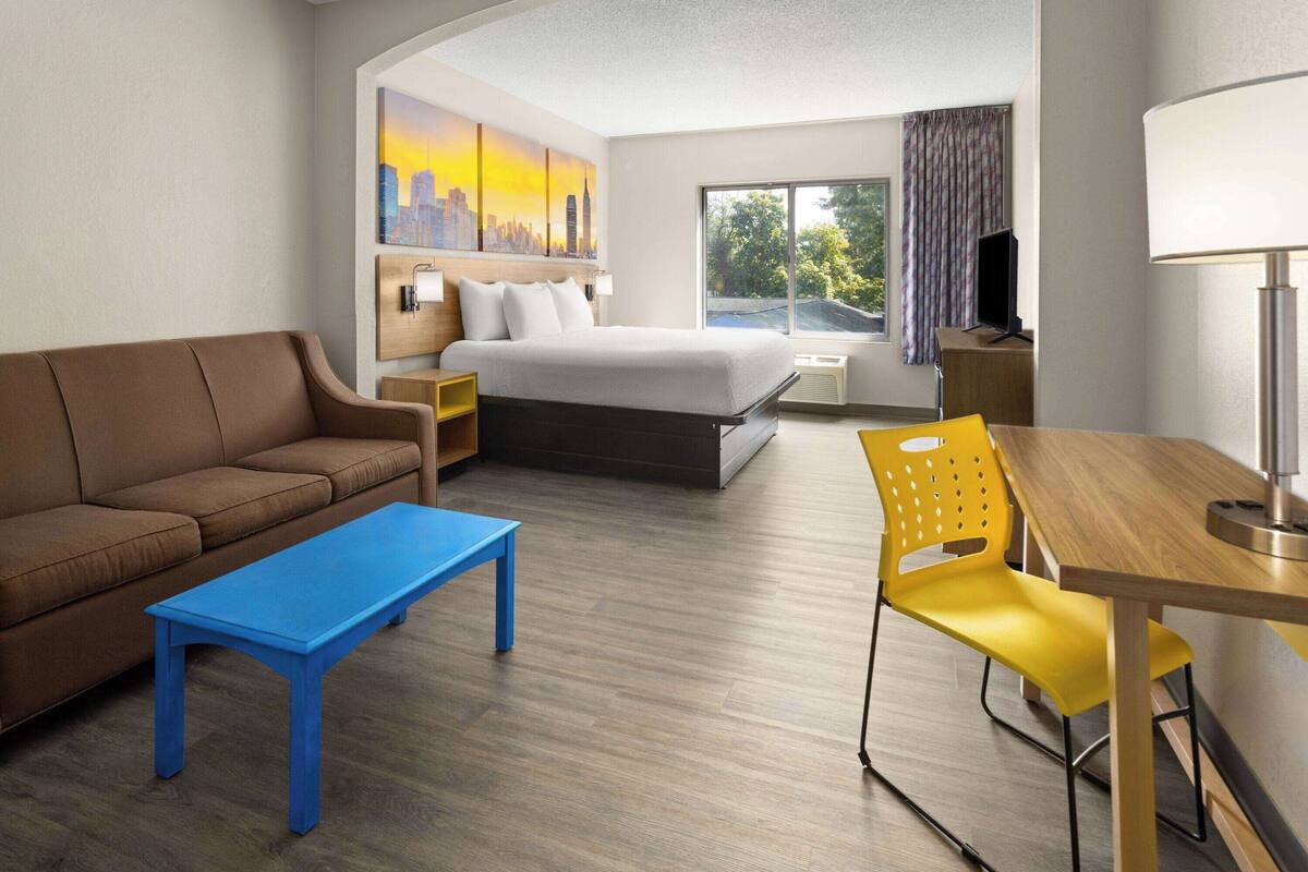Habitación de hotel con una cama, un sofá, un escritorio, una silla, un televisor y un cuadro de un paisaje urbano.