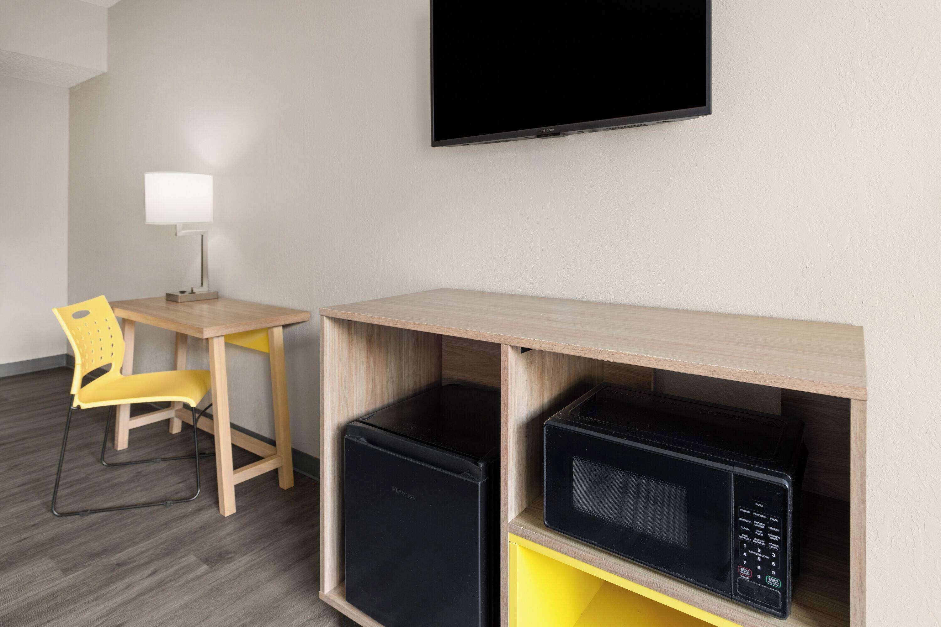 Et kompakt hotelværelse med skrivebord, stol, fladskærms-TV og indbyggede apparater, herunder et køleskab og en mikrobølgeovn.