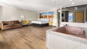 A hotel room with a wooden floor, a sofa, a bed, a desk, a lamp, a large mirror, and a bathtub.