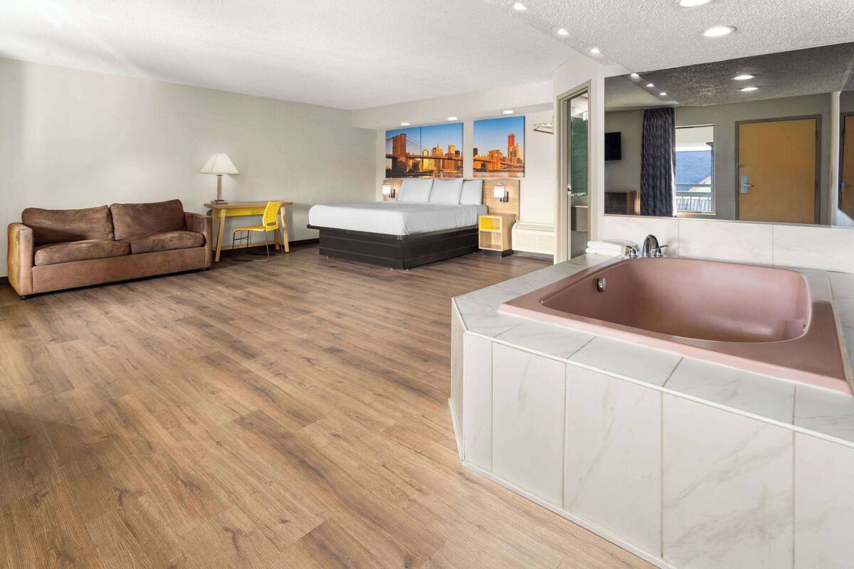 Habitación de hotel con piso de madera, un sofá, cama, escritorio, lámpara, un espejo grande y una bañera.