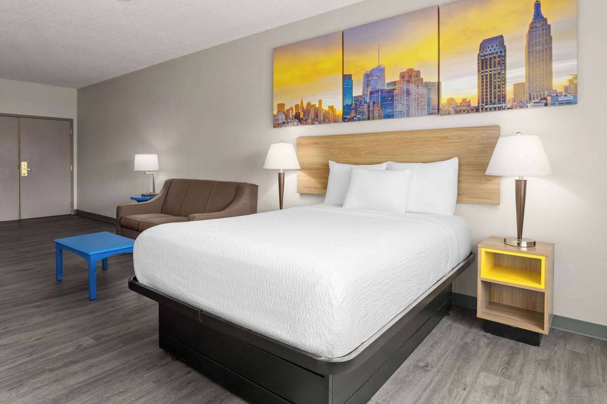 Habitación de hotel con cama, dos mesitas de noche, una silla, una mesita auxiliar azul y un cuadro de paisaje urbano en la pared.