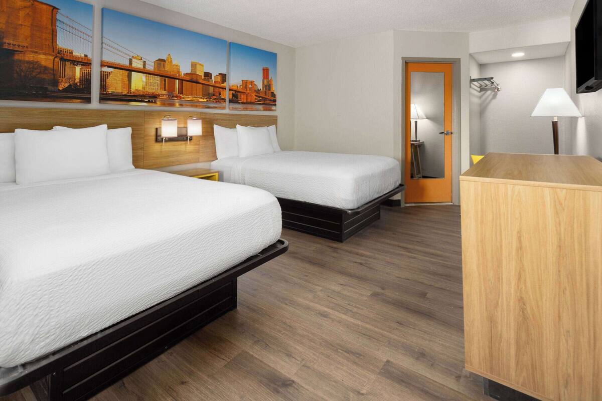 Habitación de hotel con dos camas, un escritorio de madera, una mesita de noche y una lámpara de pared.