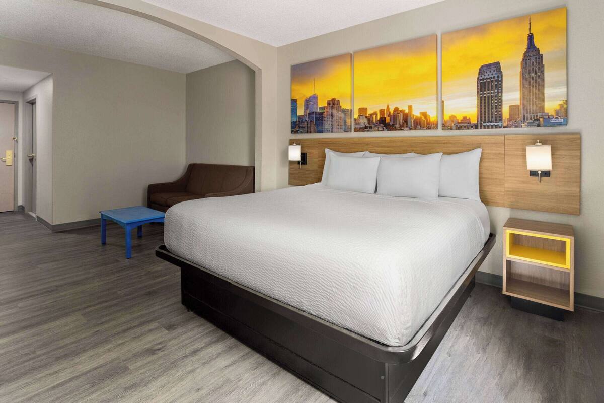 Habitación de hotel con una cama grande, una silla azul, una mesita de noche y un cuadro de un paisaje urbano en la pared.