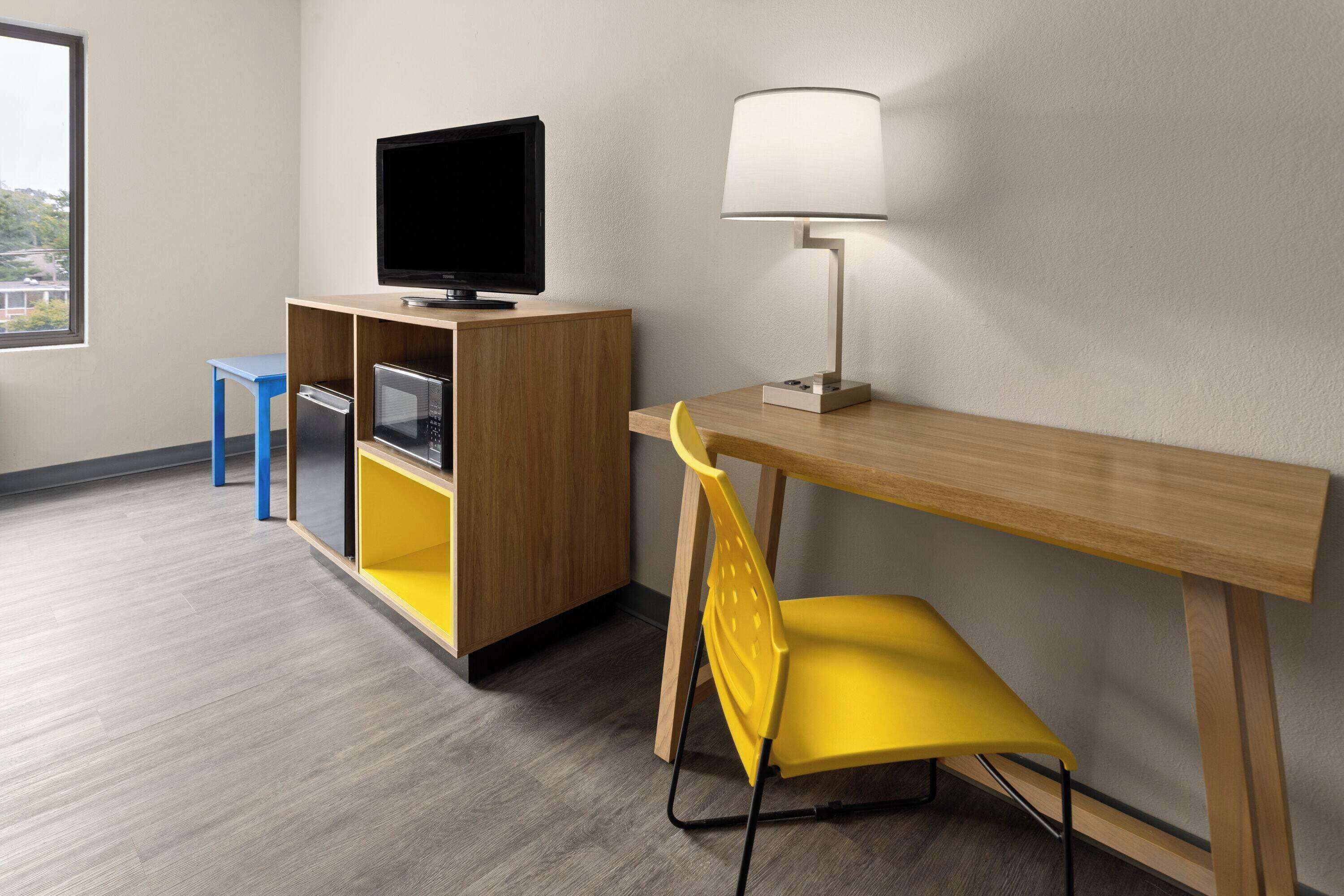 A room with a wooden desk, a yellow chair, a television on a cabinet, and a lamp.