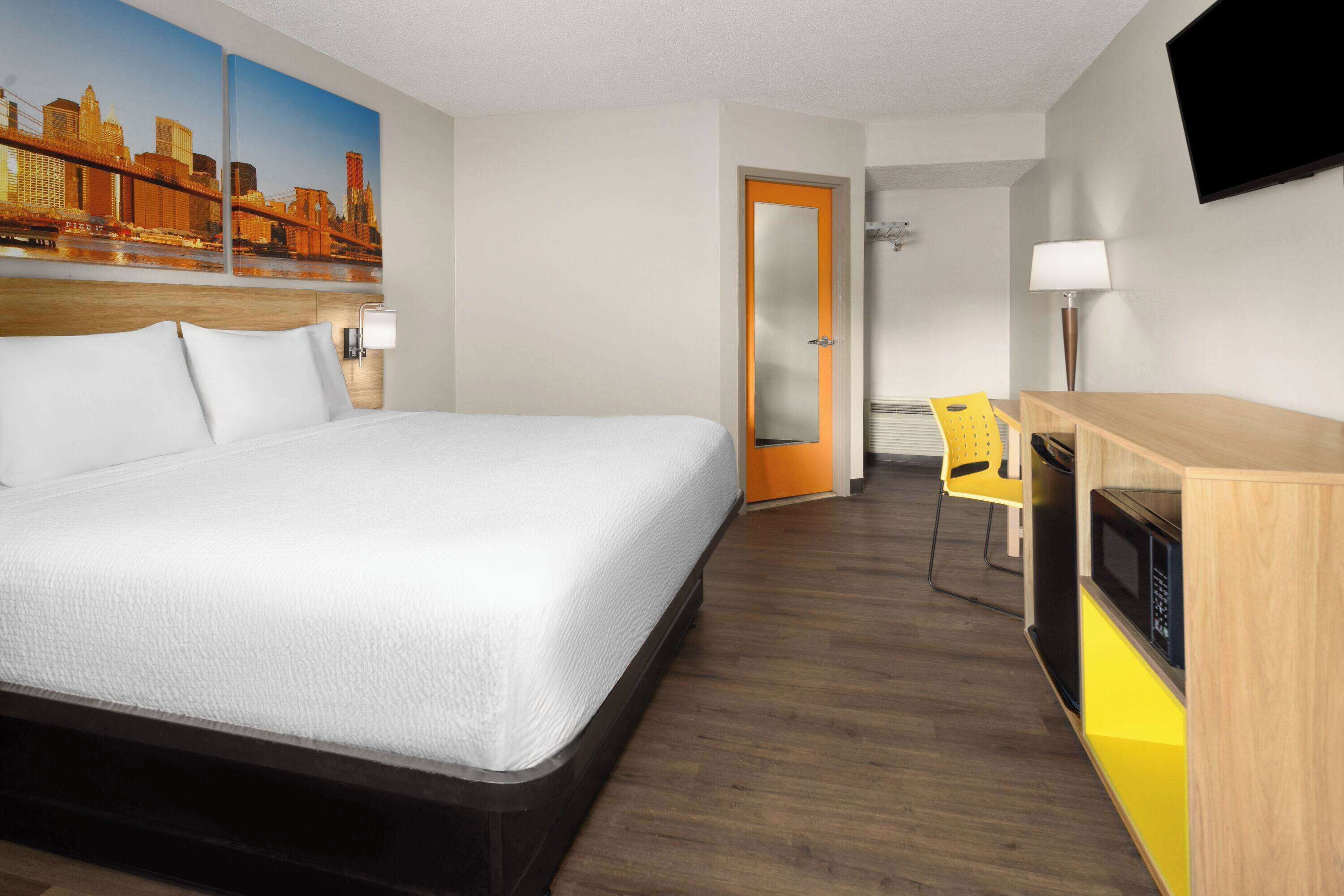 A hotel room with a large bed, a desk with a microwave, a chair, a television, and a cityscape painting on the wall.